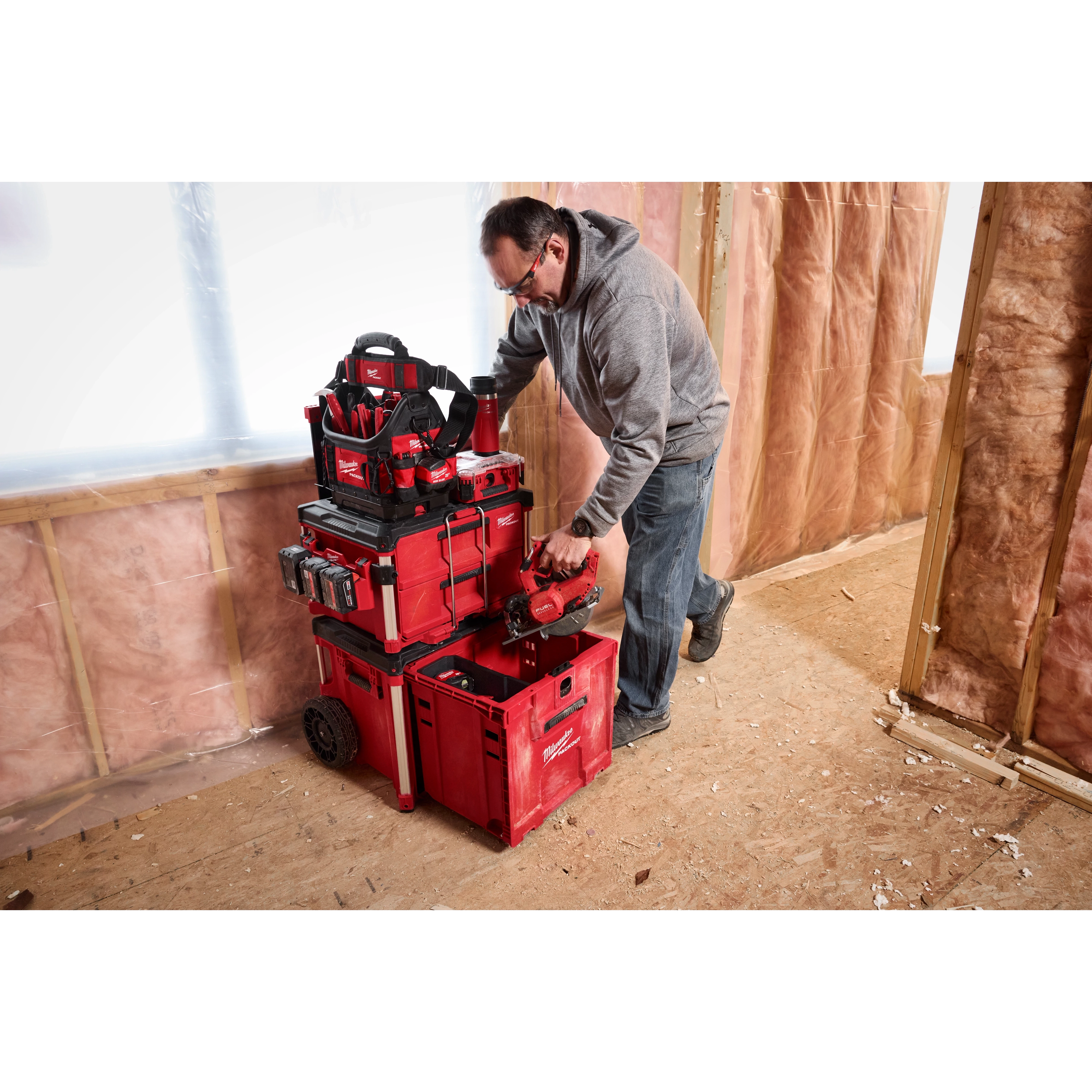A person is organizing a PACKOUT™ Rolling Drawer Tool Box system in a construction area with pink insulation on the walls. The red modular toolbox system features stacked storage compartments and various tools.
