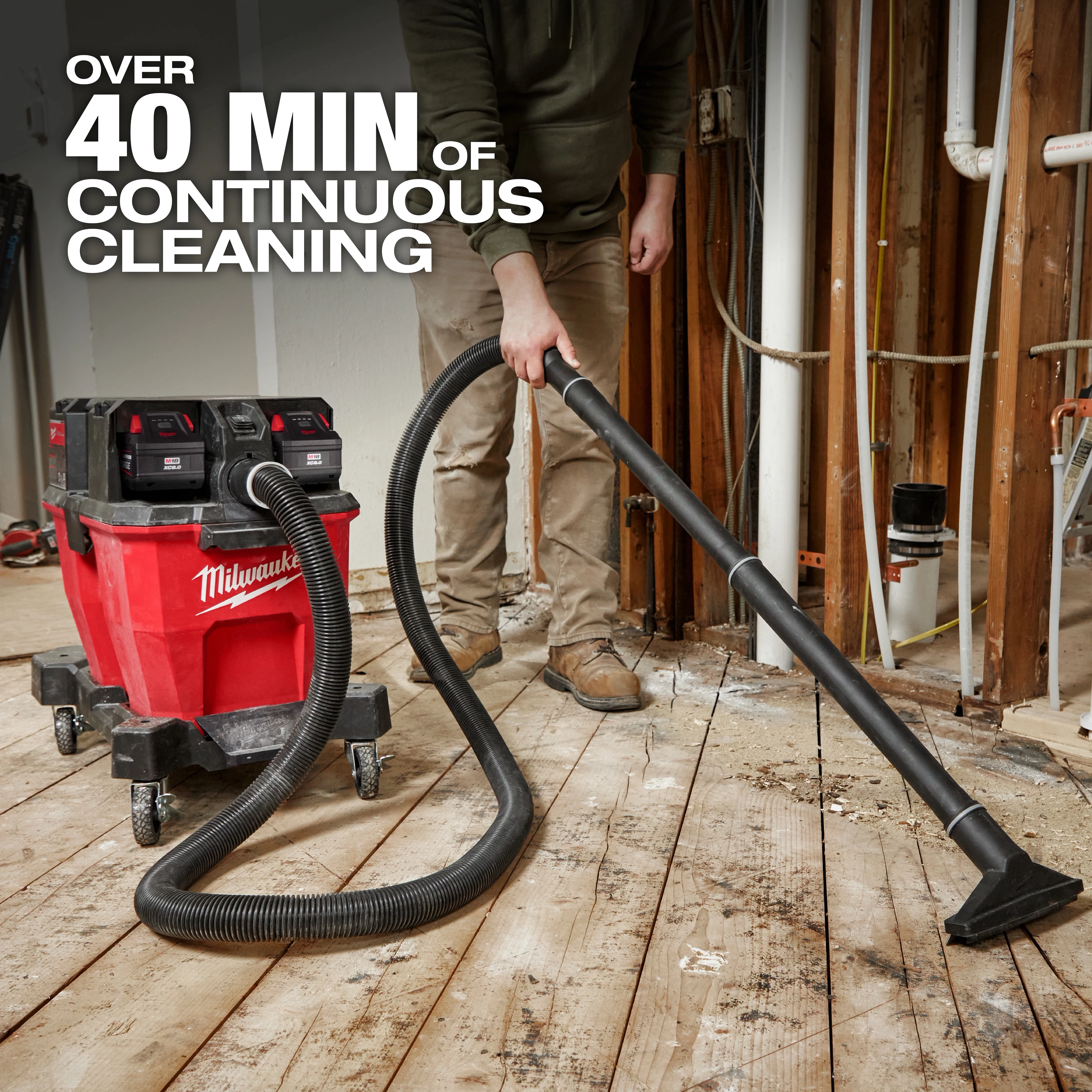 The M18 FUEL™ NEXUS™ 9 Gallon Dual-Battery Wet/Dry Vacuum offers over 40 minutes of continuous cleaning. It features a sturdy red canister on wheels with black dual battery compartments. A person uses the attached hose to clean debris from a wooden floor in a construction site setting.
