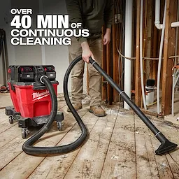 The M18 FUEL™ NEXUS™ 9 Gallon Dual-Battery Wet/Dry Vacuum offers over 40 minutes of continuous cleaning. It features a sturdy red canister on wheels with black dual battery compartments. A person uses the attached hose to clean debris from a wooden floor in a construction site setting.