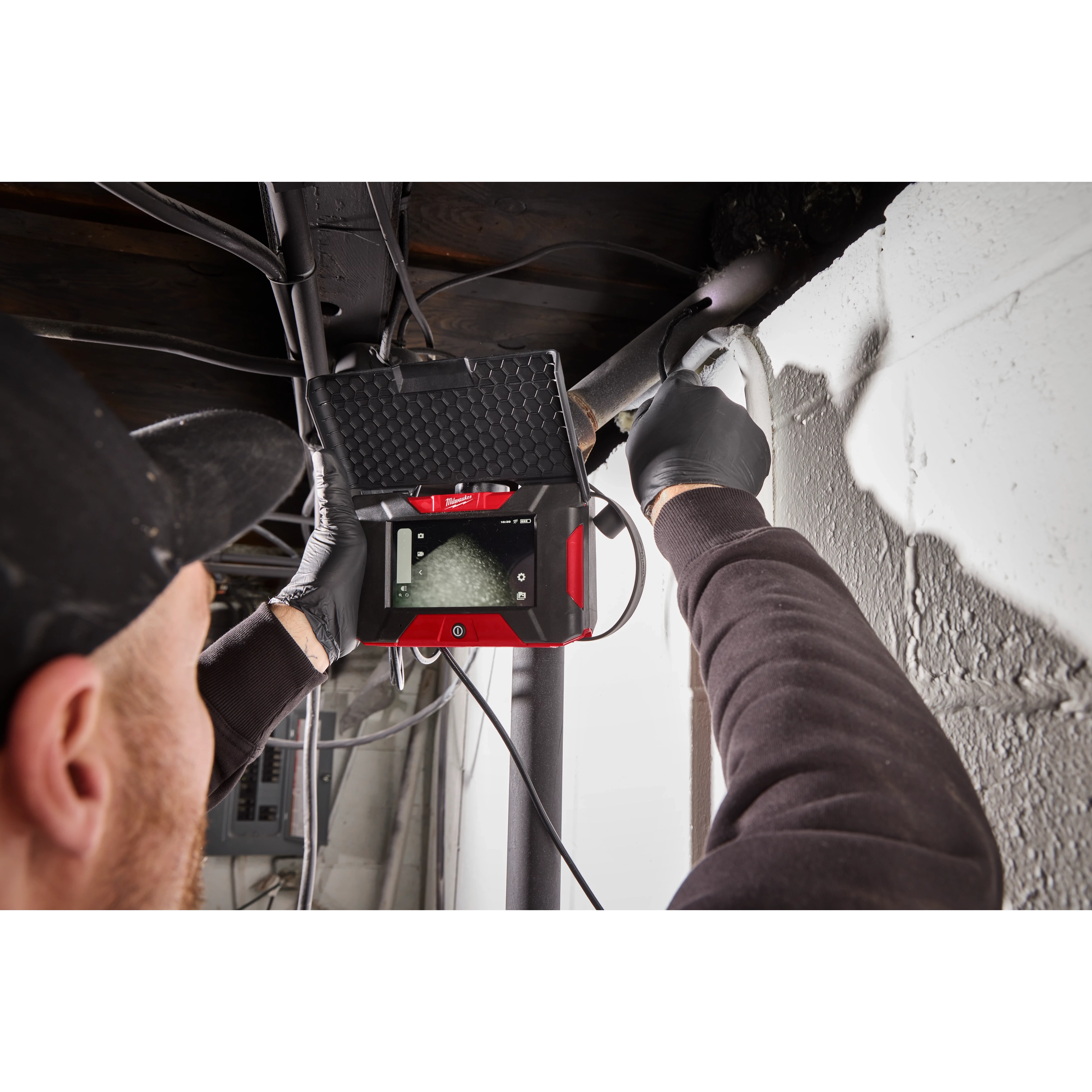 A person is using an M12™ Wireless Monitor to inspect a pipe near a wall. The device has a red and black casing and is displaying the interior view of the pipe on its screen. The individual is wearing gloves and a black cap.