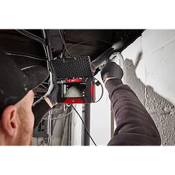 A person is using an M12™ Wireless Monitor to inspect a pipe near a wall. The device has a red and black casing and is displaying the interior view of the pipe on its screen. The individual is wearing gloves and a black cap.