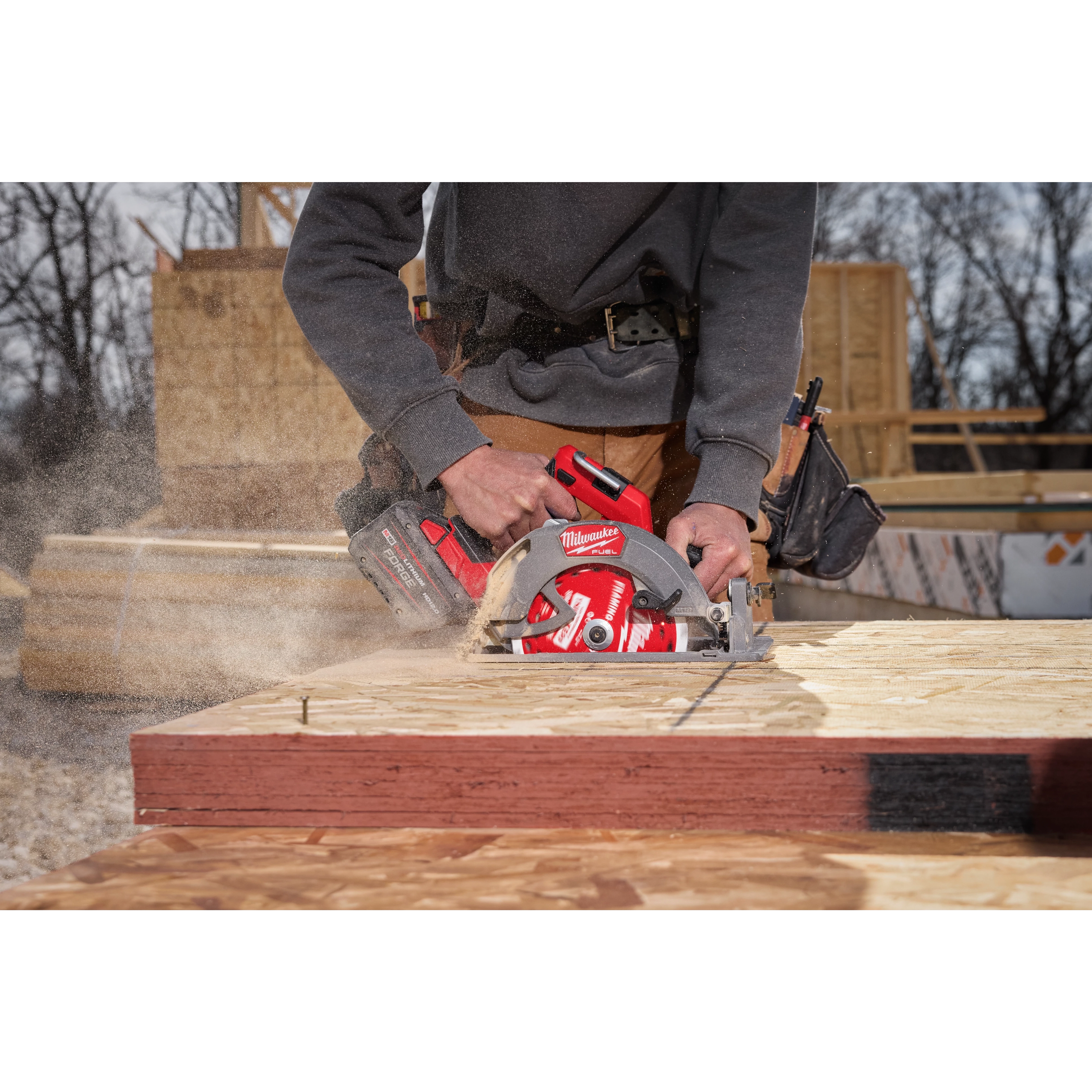 A worker uses an M18 FUEL™ 7-1/4” Circular Saw to cut through a piece of wood on an outdoor construction site. Sawdust is visible coming from the cutting area, and the worker is wearing a gray sweatshirt and brown work pants. Various construction materials are in the background.