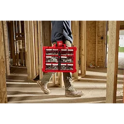 A person in work boots and green pants carries a red plastic organizer box filled with various small tools and hardware. The individual is walking through a wooden framed construction site, with partially completed walls and beams visible. Bright sunlight streams through the space.