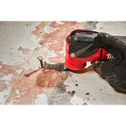 A worker using a MILWAUKEE® OPEN-LOK™ 1-3/8" Carbide Teeth Metal Multi-Tool Blade to cut through an exposed nail on a worn floor. The hand and tool are focused in the foreground, demonstrating the blade's precision in cutting metal.