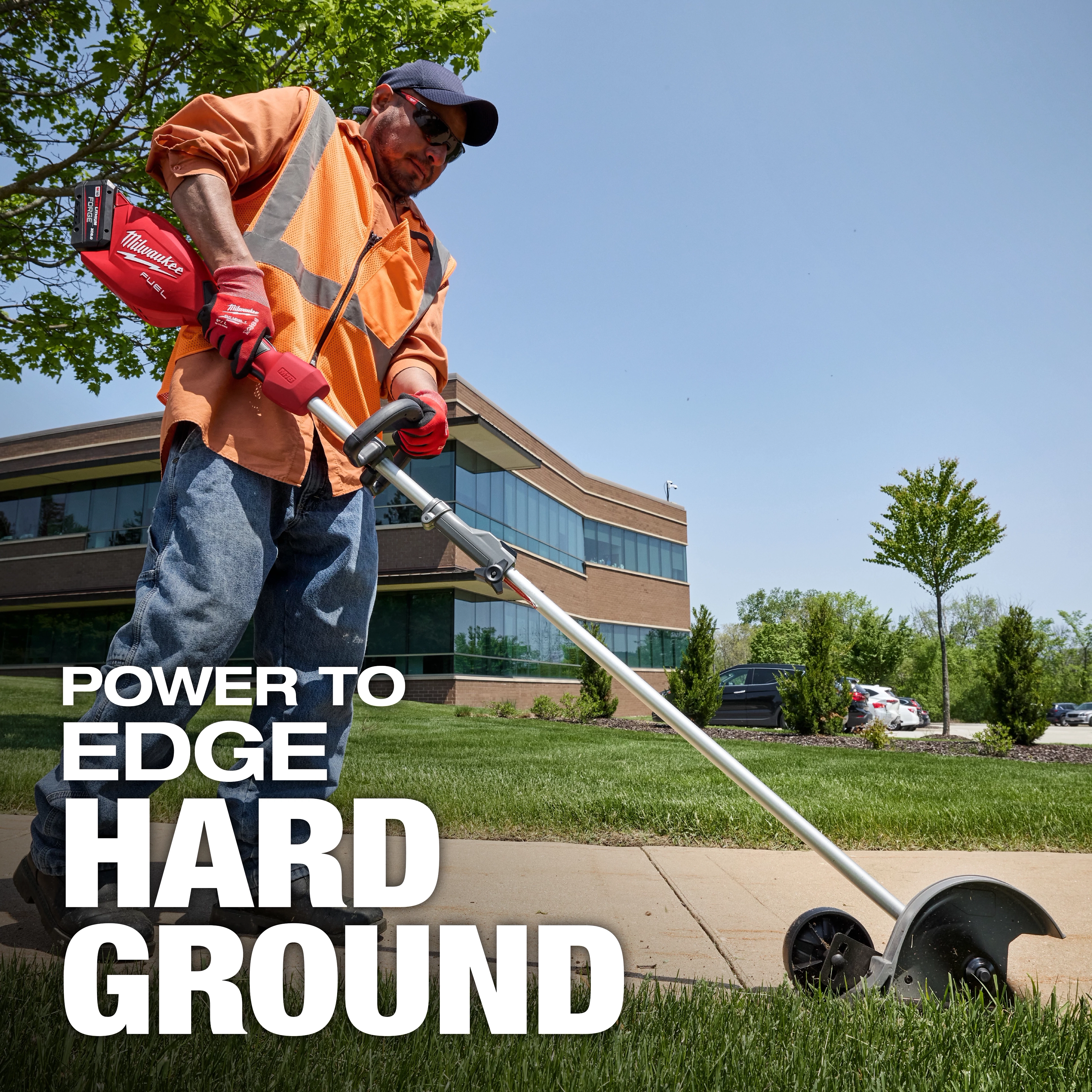 A worker in an orange vest and jeans uses a Milwaukee power tool to edge the grass along a sidewalk. The background shows a modern office building, green trees, and a clear blue sky. The text on the image reads, "POWER TO EDGE HARD GROUND."