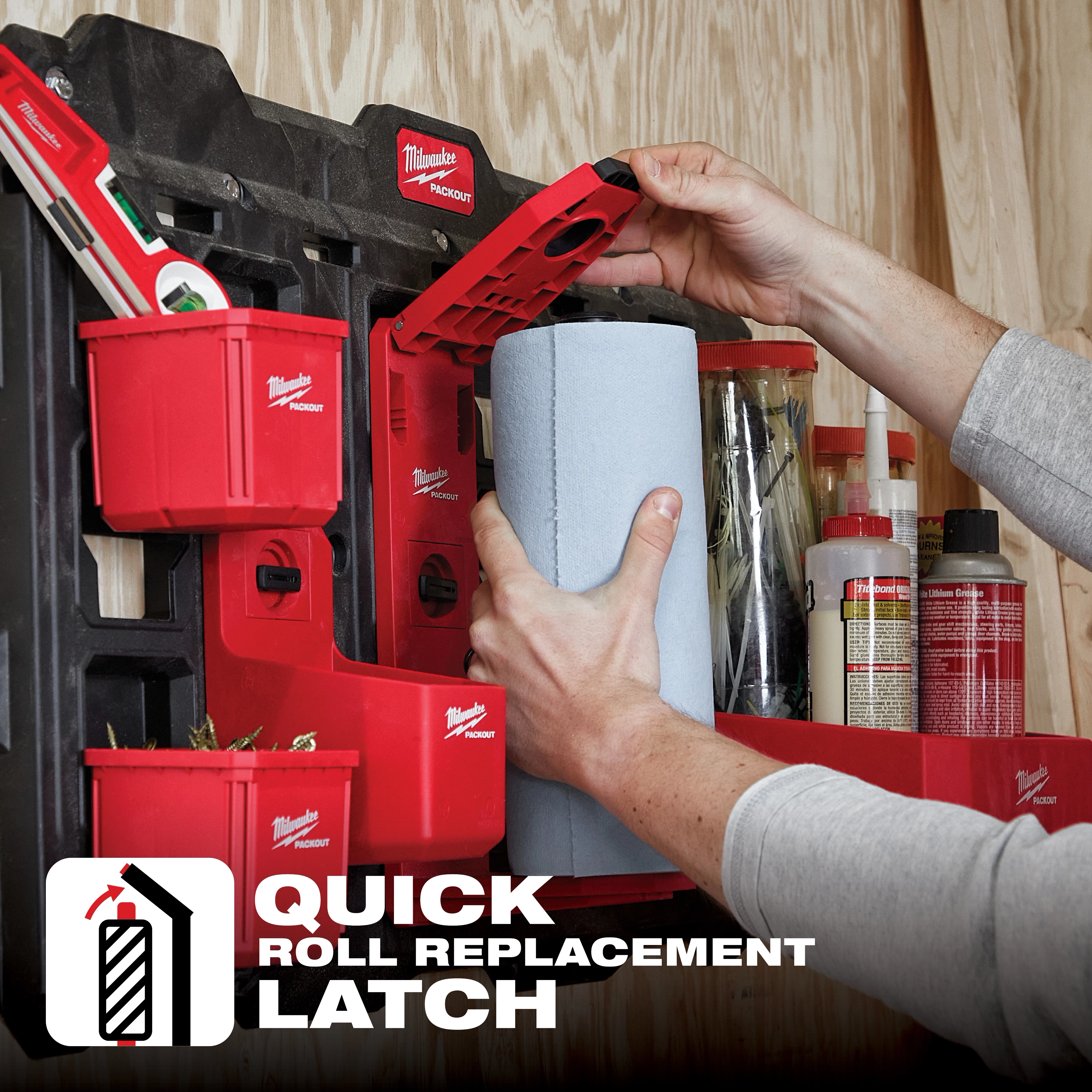 A person installing a blue paper roll onto a red PACKOUT Roll Holder attached to a wall with additional storage compartments.