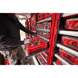 The PACKOUT™ Rack Kit is shown in use. A person in workwear retrieves a red, compartmentalized toolbox from the rack system installed in a work van. The rack organizes multiple red and gray toolboxes efficiently against the walls.
