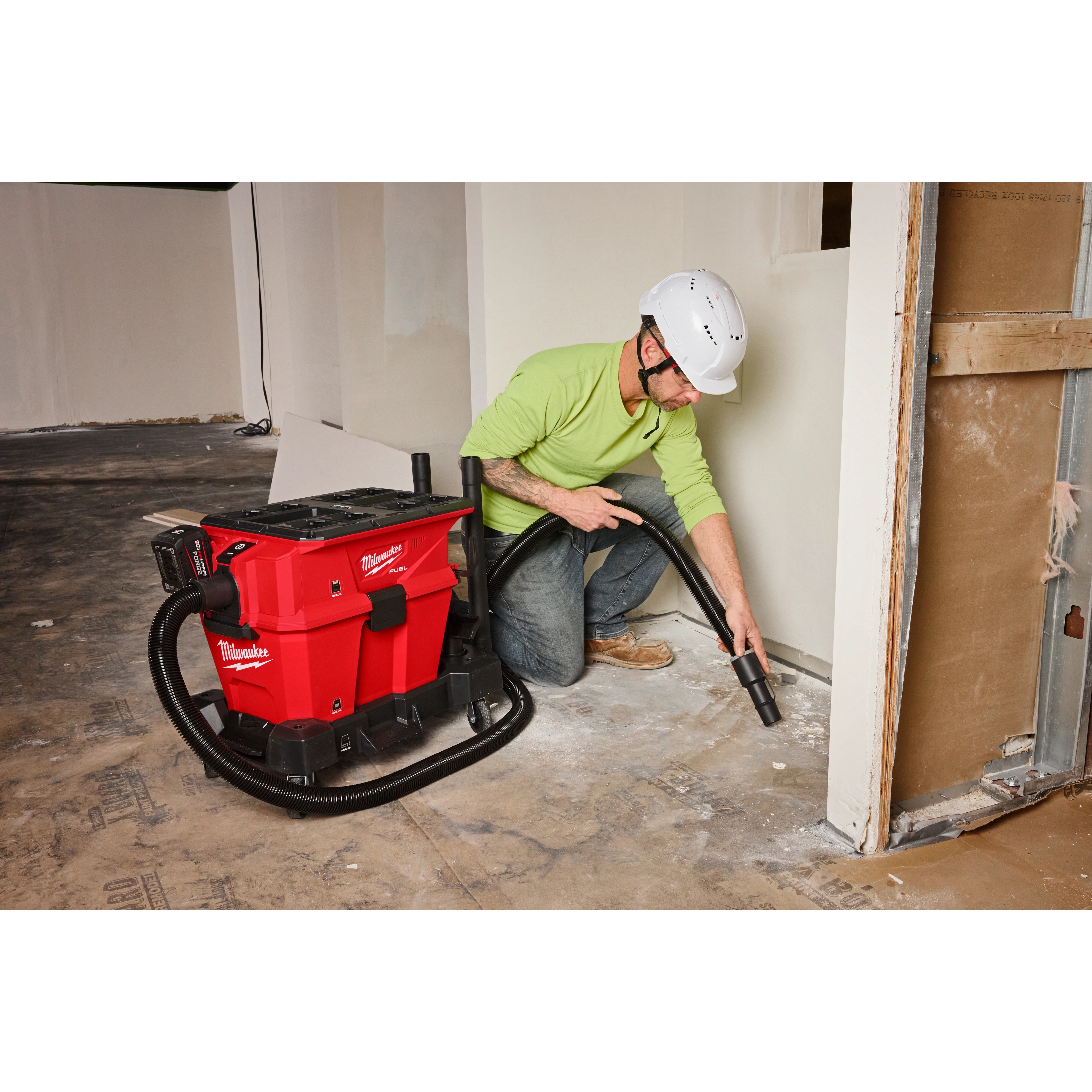 A worker uses a Milwaukee M18 cordless wet/dry vacuum to clean a construction site. The vacuum is red with black accents and has a long hose. The worker wears a lime green shirt, blue jeans, brown shoes, and a white hard hat.