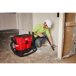 A worker uses a Milwaukee M18 cordless wet/dry vacuum to clean a construction site. The vacuum is red with black accents and has a long hose. The worker wears a lime green shirt, blue jeans, brown shoes, and a white hard hat.