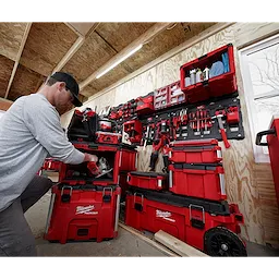A person uses a PACKOUT™ Cabinet, which is part of a red modular storage system filled with various tools. The setup is mounted on a wooden wall within a work shed, with additional storage units organized efficiently on and around the cabinet.