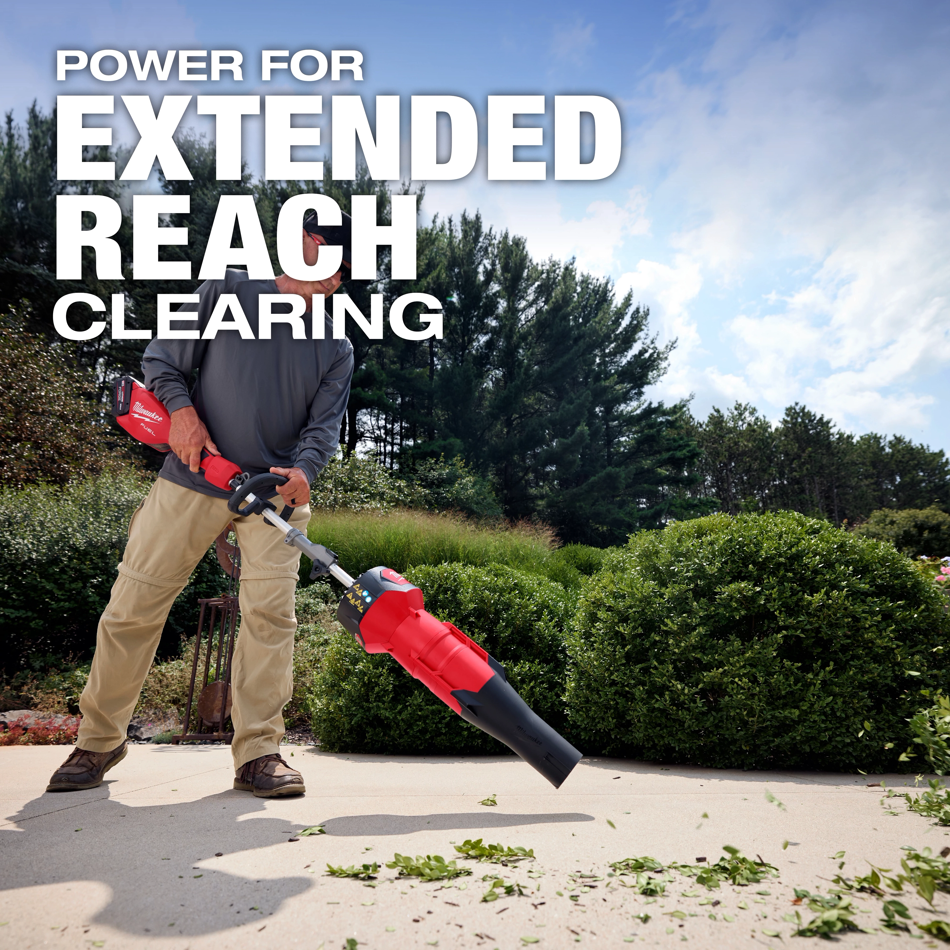 A man uses a handheld leaf blower to clear debris from a sidewalk. The text overlay reads "POWER FOR EXTENDED REACH CLEARING." Behind him are lush green bushes and tall trees under a partly cloudy sky.