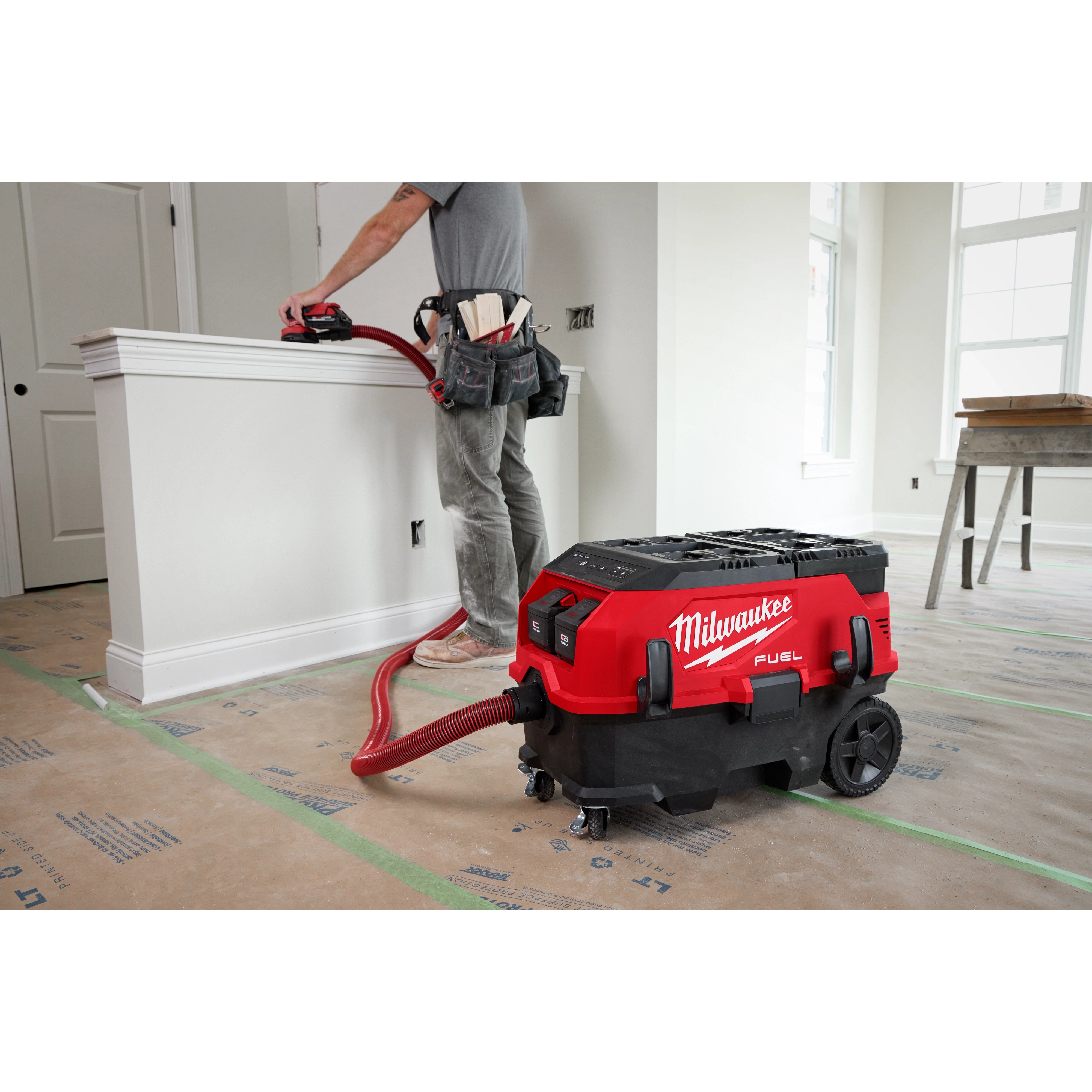 A person is using a red, M18 FUEL™ PACKOUT™ 9 Gallon Dual-Battery Dust Extractor with VACLINK™. The extractor is connected by a hose and is on a construction site, with protective covering on the floor.