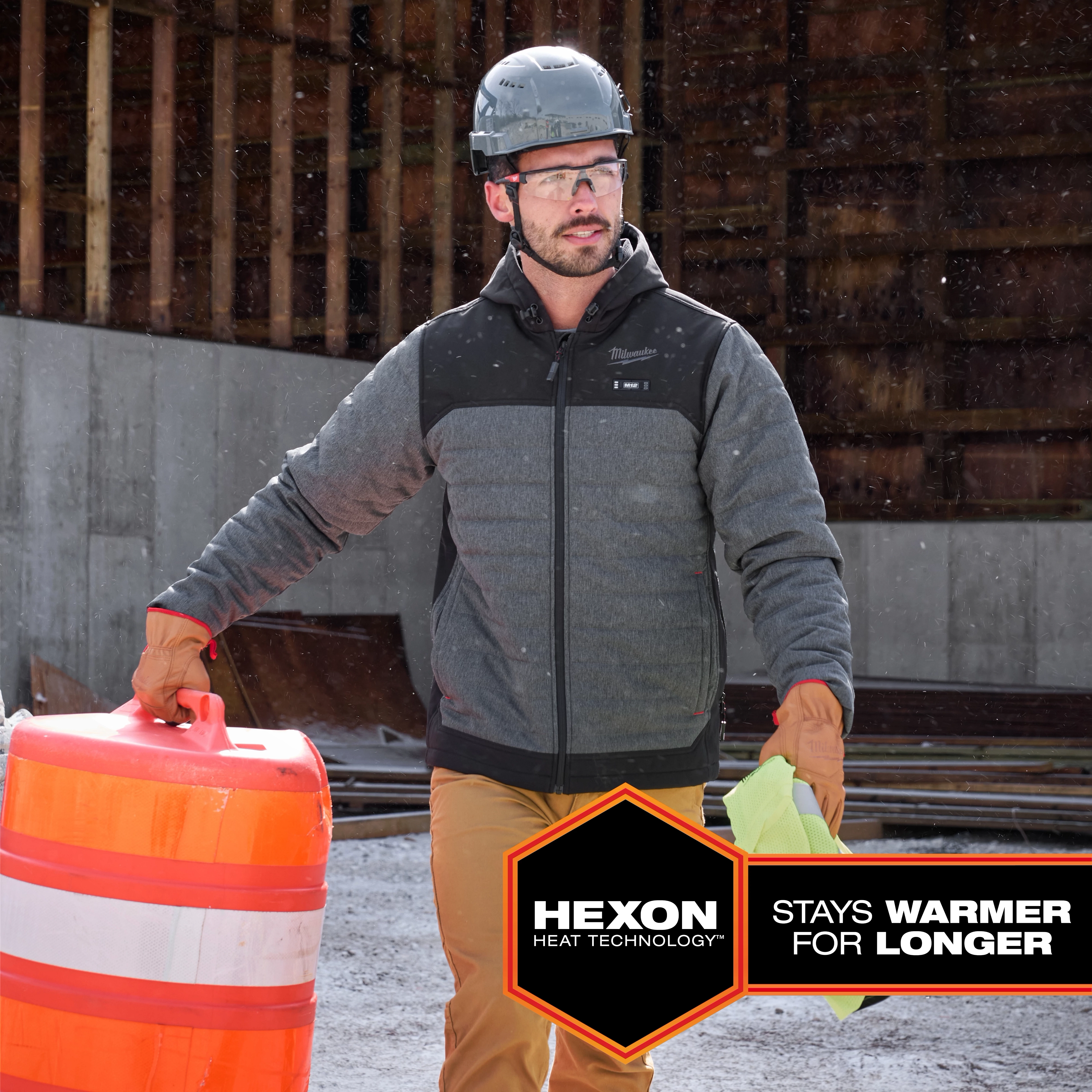 Person wearing the M12 Heated AXIS Jacket at a construction site, carrying an orange-and-white traffic barrel and a yellow cloth.