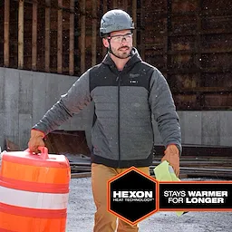 Person wearing the M12 Heated AXIS Jacket at a construction site, carrying an orange-and-white traffic barrel and a yellow cloth.