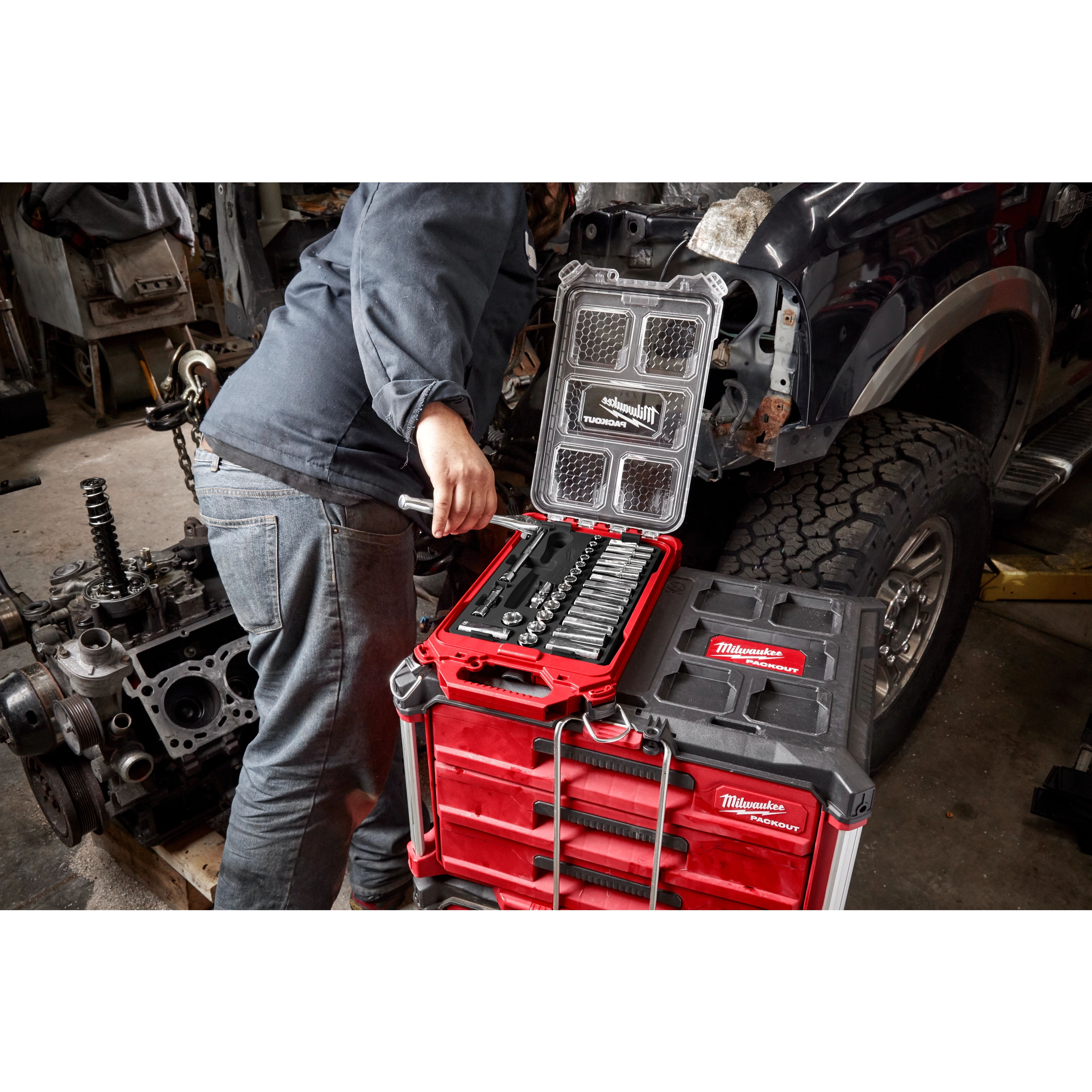 A person works on a vehicle in a garage, with various tools and equipment around. They are using a toolset from a Milwaukee Packout box placed on a red Milwaukee rolling toolbox. An engine block is visible on the left side of the image.