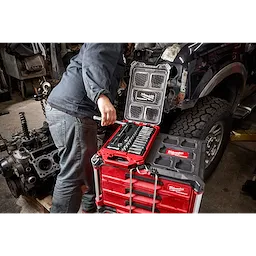 A person works on a vehicle in a garage, with various tools and equipment around. They are using a toolset from a Milwaukee Packout box placed on a red Milwaukee rolling toolbox. An engine block is visible on the left side of the image.