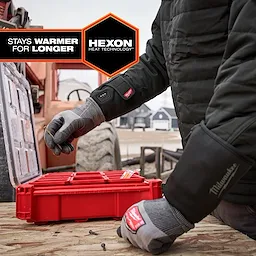 A person wearing Milwaukee Heated Work Glove Liners organizes items in a red storage box outdoors. The gloves feature Hexon Heat Technology and promise to stay warmer for longer. A construction site and large machinery are visible in the background. The gloves have black cuffs.