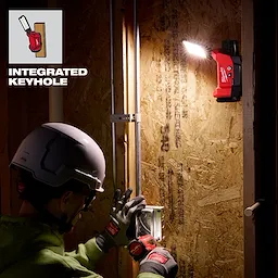 M12™ ROVER™ Multi-Directional Flood Light w/ PACKOUT™ Compatibility illuminates a work area as a person wearing a hard hat and gloves installs an electrical outlet. The flood light is mounted on a wooden panel via an integrated keyhole. The light beam highlights the working space efficiently.