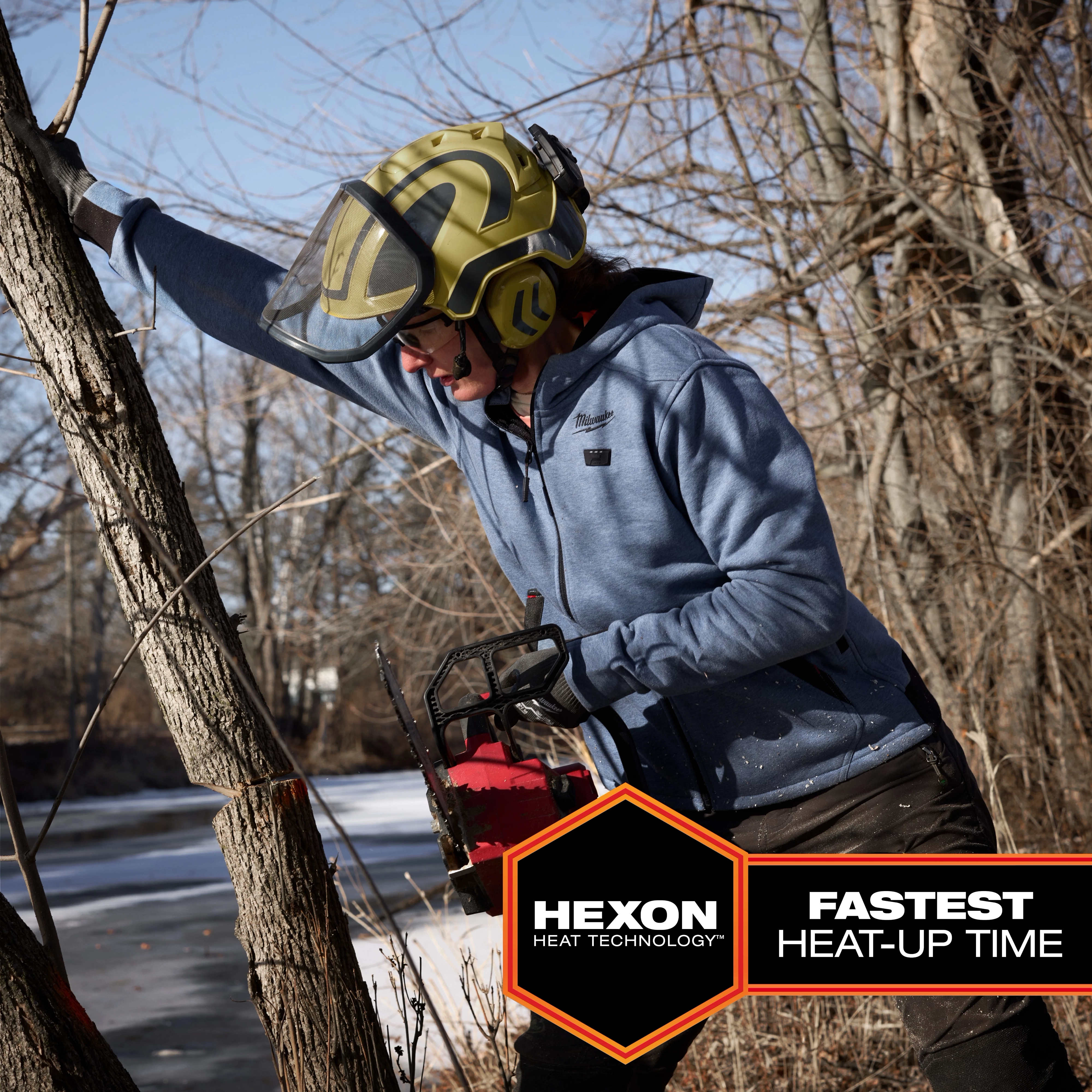 The M12™ Heated Hoodie is shown being worn by a person using a chainsaw outdoors in a wooded area. The hoodie features HEXON heat technology and provides the fastest heat-up time, depicted in text on the image. It is blue and has a front zipper and pockets.