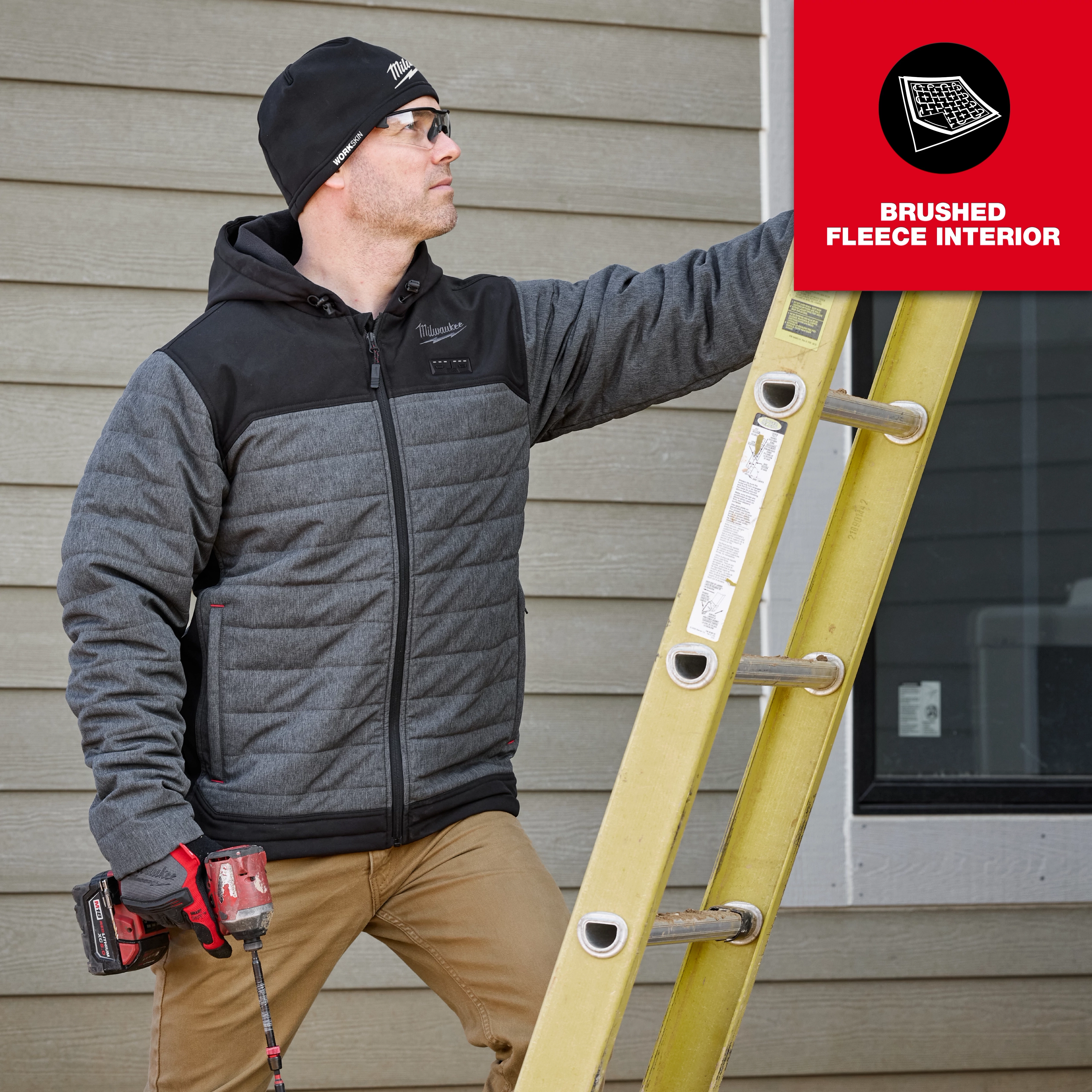 A person wearing a WORKSKIN™ Low-Profile Beanie, gray and black zip-up jacket, and brown pants is holding a red power tool and climbing a yellow ladder. A red label in the top right corner reads "BRUSHED FLEECE INTERIOR."