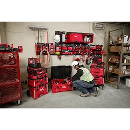 A worker in a hard hat and safety vest organizes tools with the PACKOUT™ Shop Storage Breadth of Line Image system, comprising red storage boxes and wall-mounted units filled with various tools and accessories. The system is designed for efficient organization on a shop floor.