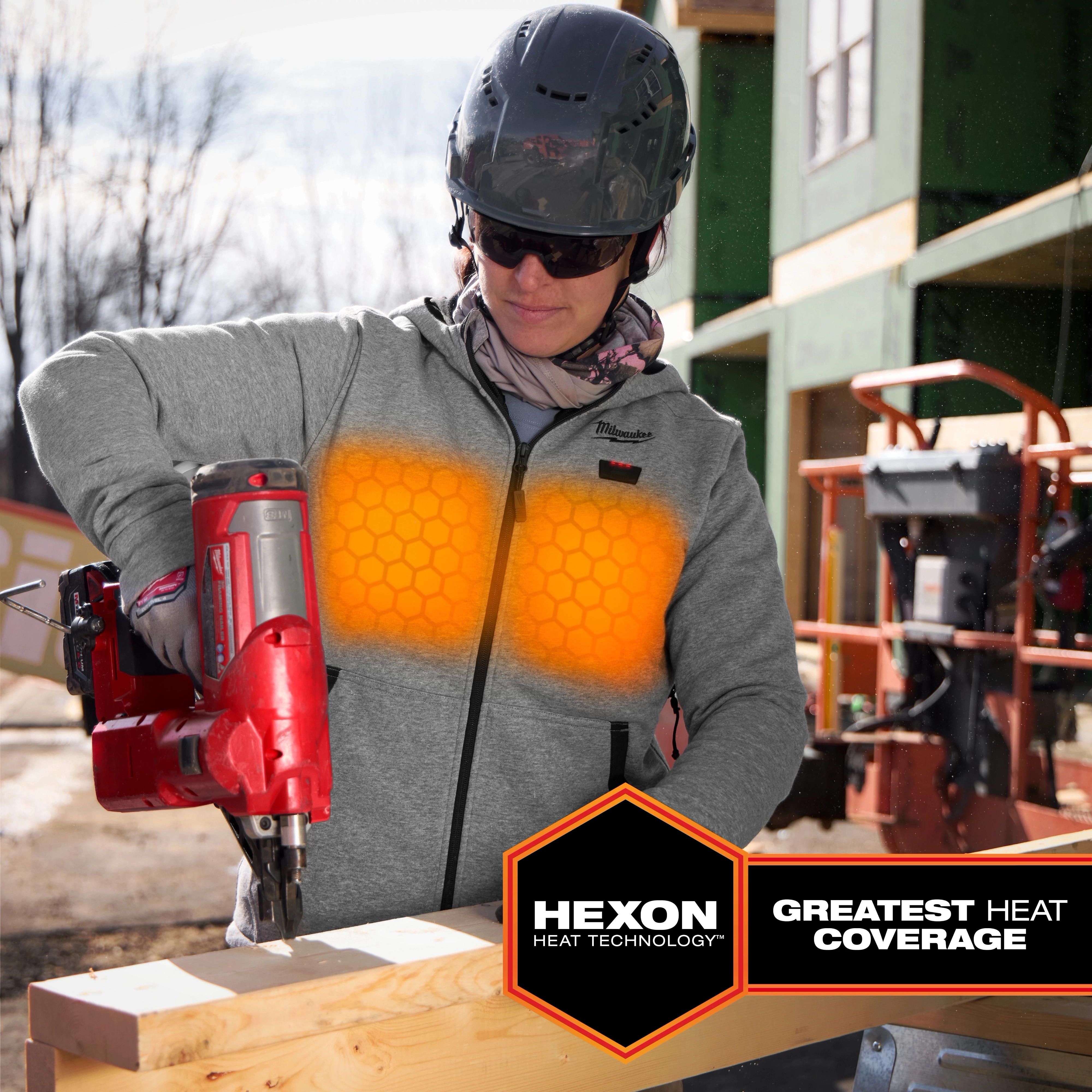 A construction worker uses a tool while wearing the M12™ Heated Hoodie, which features HEXON Heat Technology for the greatest heat coverage. The hoodie is gray with a hexagonal pattern glowing orange to indicate its heating function. The worker wears a helmet and stands in front of a construction site.