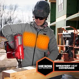 A construction worker uses a tool while wearing the M12™ Heated Hoodie, which features HEXON Heat Technology for the greatest heat coverage. The hoodie is gray with a hexagonal pattern glowing orange to indicate its heating function. The worker wears a helmet and stands in front of a construction site.