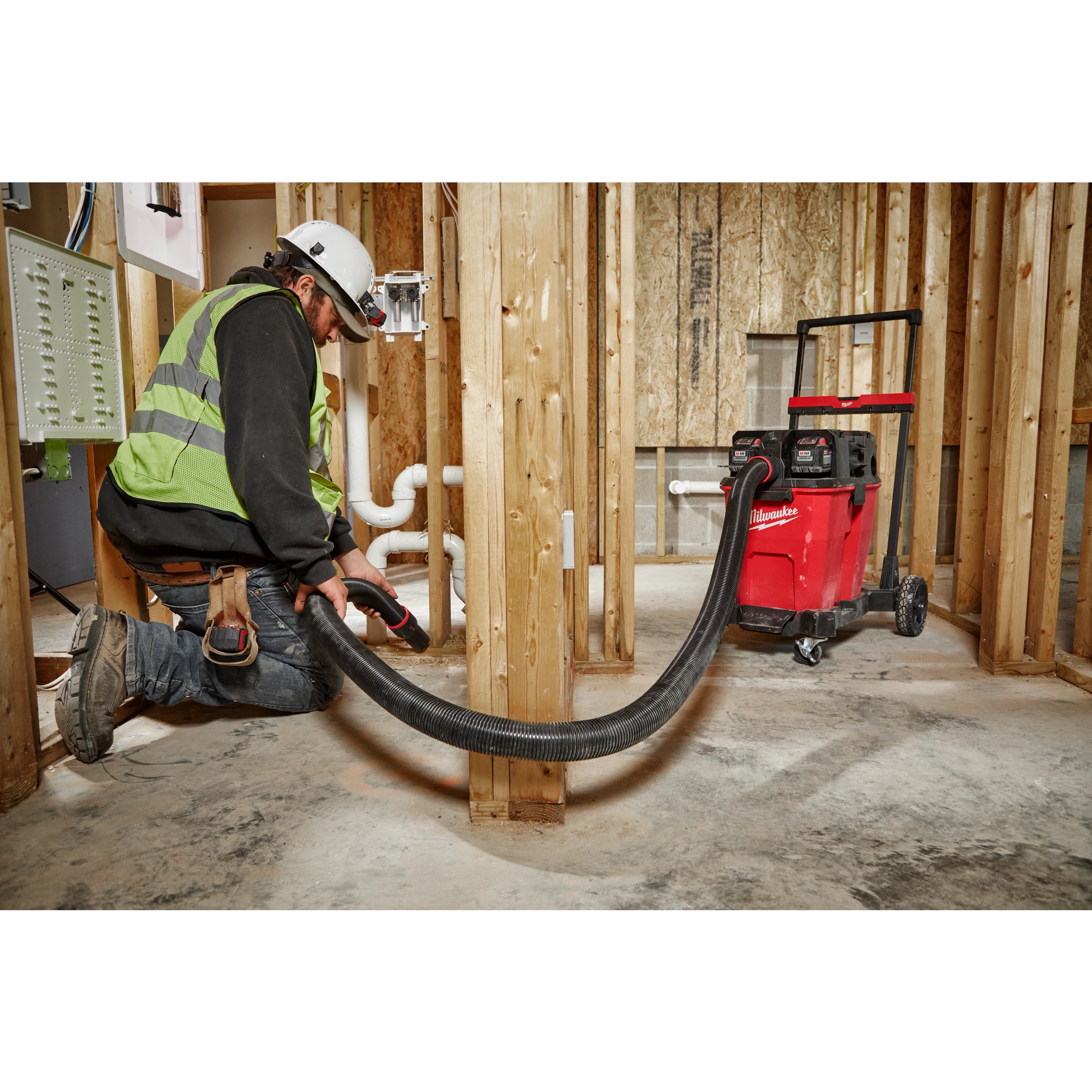 Worker using a 2-1/2” x 9' Flexible Hose attached to a red vacuum in a construction setting with exposed wooden framing and pipes.