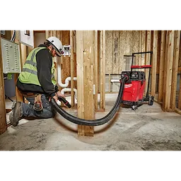 Worker using a 2-1/2” x 9' Flexible Hose attached to a red vacuum in a construction setting with exposed wooden framing and pipes.