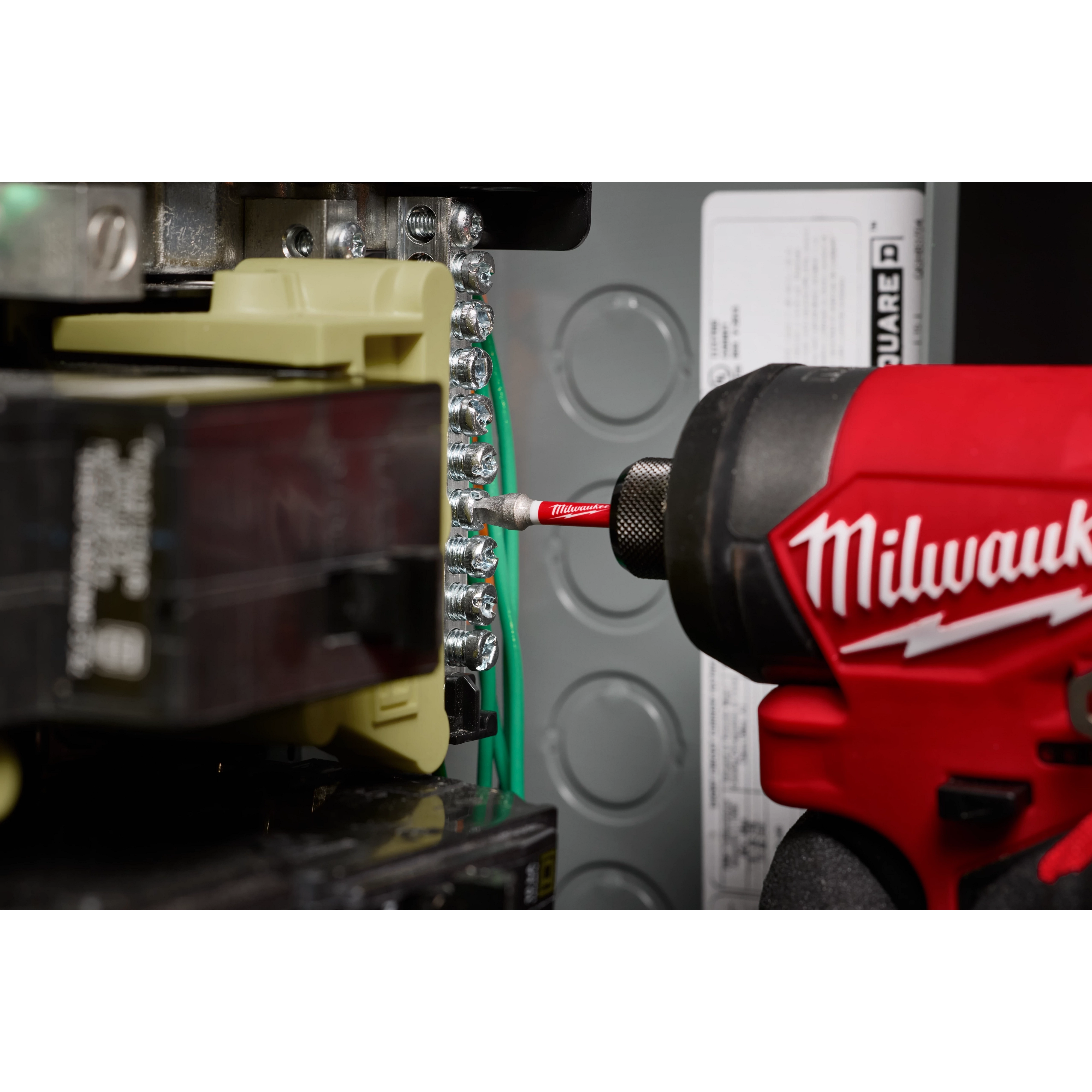 A Milwaukee power tool is being used with a SHOCKWAVE™ 2” Impact Square Recess #2 Power Bit to tighten screws on an electrical panel. The bit is attached to the power tool and is making contact with the screw head. Various wiring components are visible in the background.