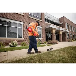 Worker with trimmer edges lawn near brick building with large windows and canopy, wearing orange shirt and blue pants.