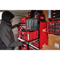 A person uses a PACKOUT™ Rack Kit by Milwaukee, organizing various red and black toolboxes on a mounted shelving unit in a workshop or garage. The rack neatly arranges all the toolboxes and other equipment.