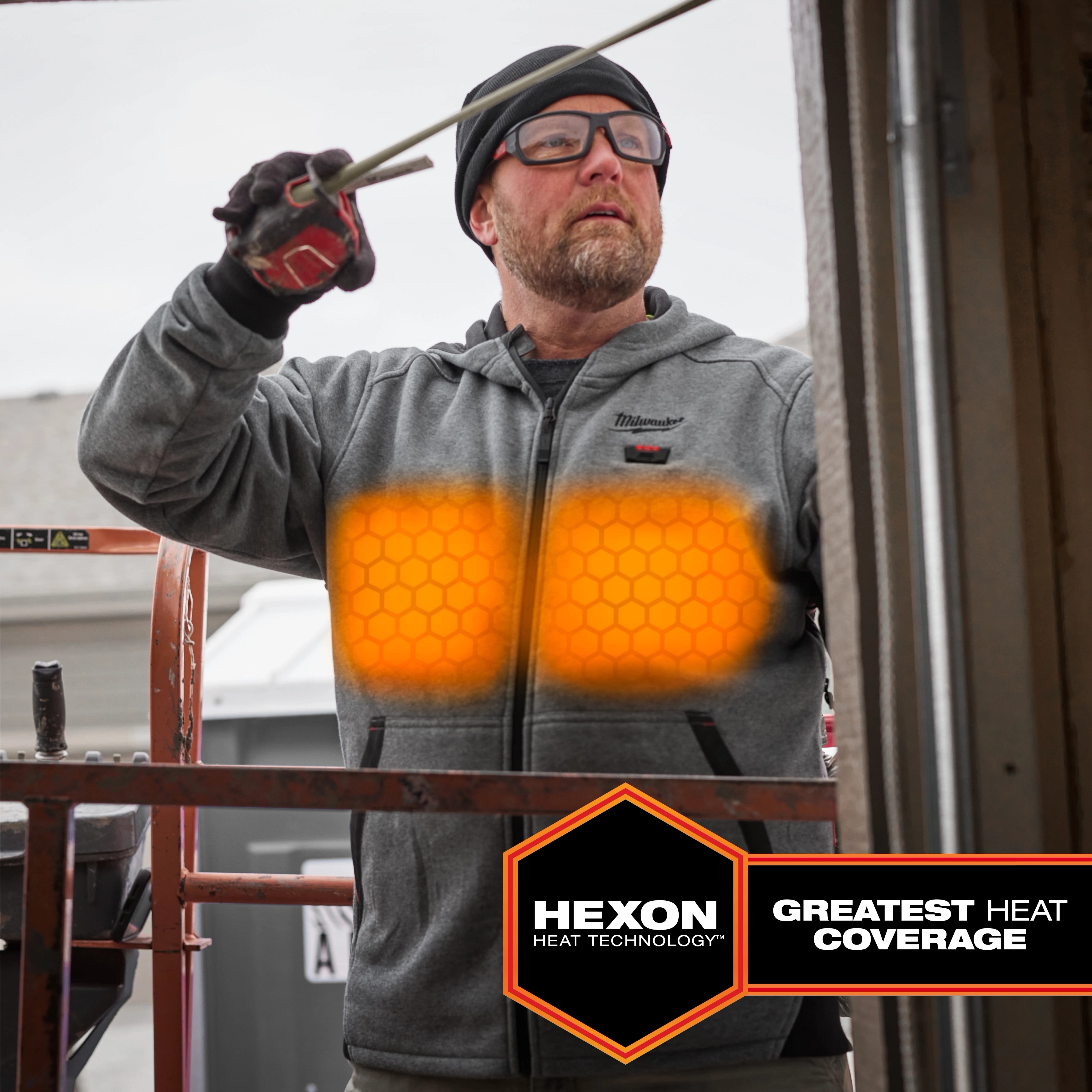 M12™ Heated Hoodie features Hexon Heat Technology for maximum heat coverage, shown with glowing heat zones on the chest. The grey hoodie has a front zipper, two pockets, and the Milwaukee logo on the upper left side. The man's gloves are visible as he works with a tool.