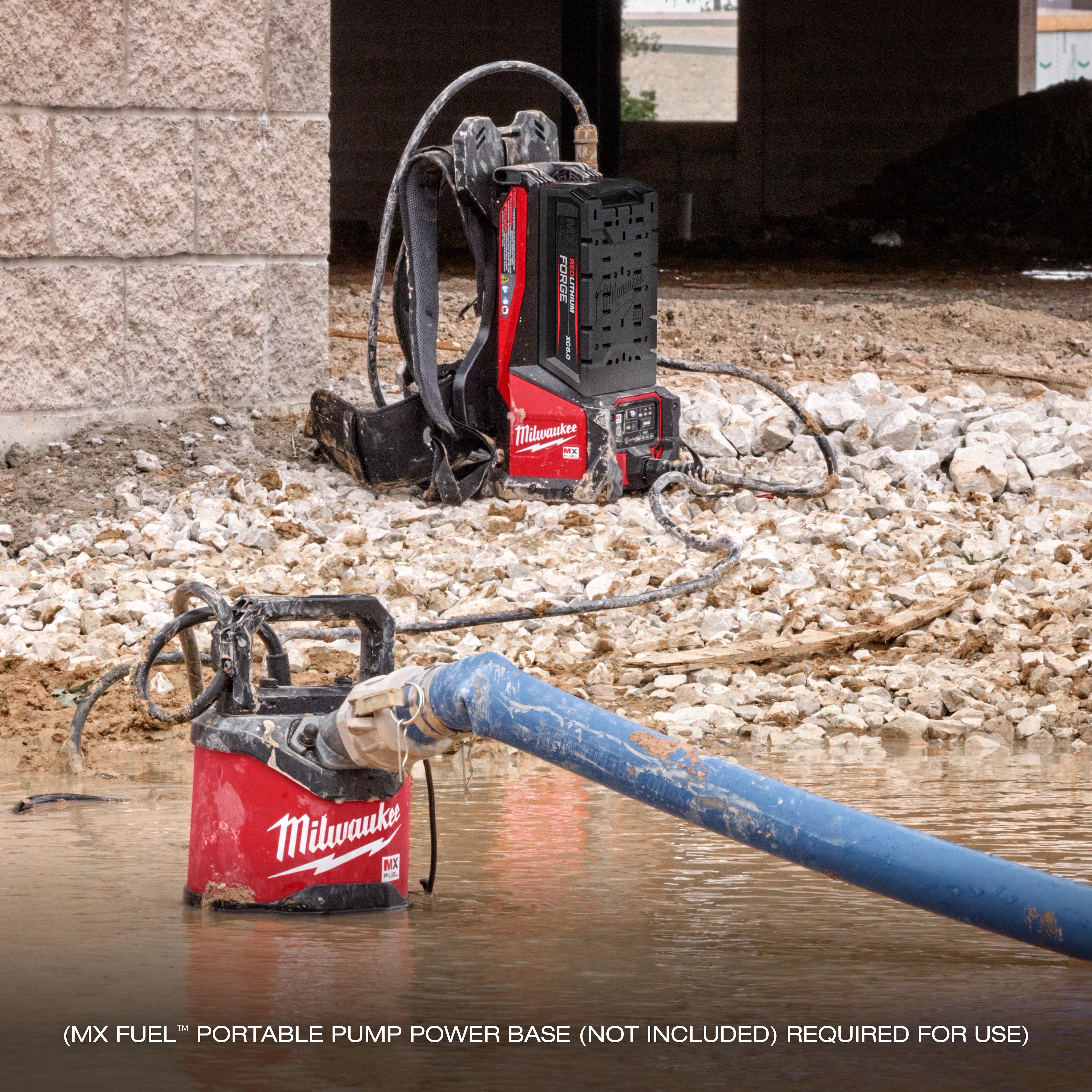 The image shows an MX FUEL™ 1HP 2" Submersible Pump in a construction setting, with the pump partially submerged in water and connected to a blue hose. In the background, there is a red and black portable pump power base (not included).