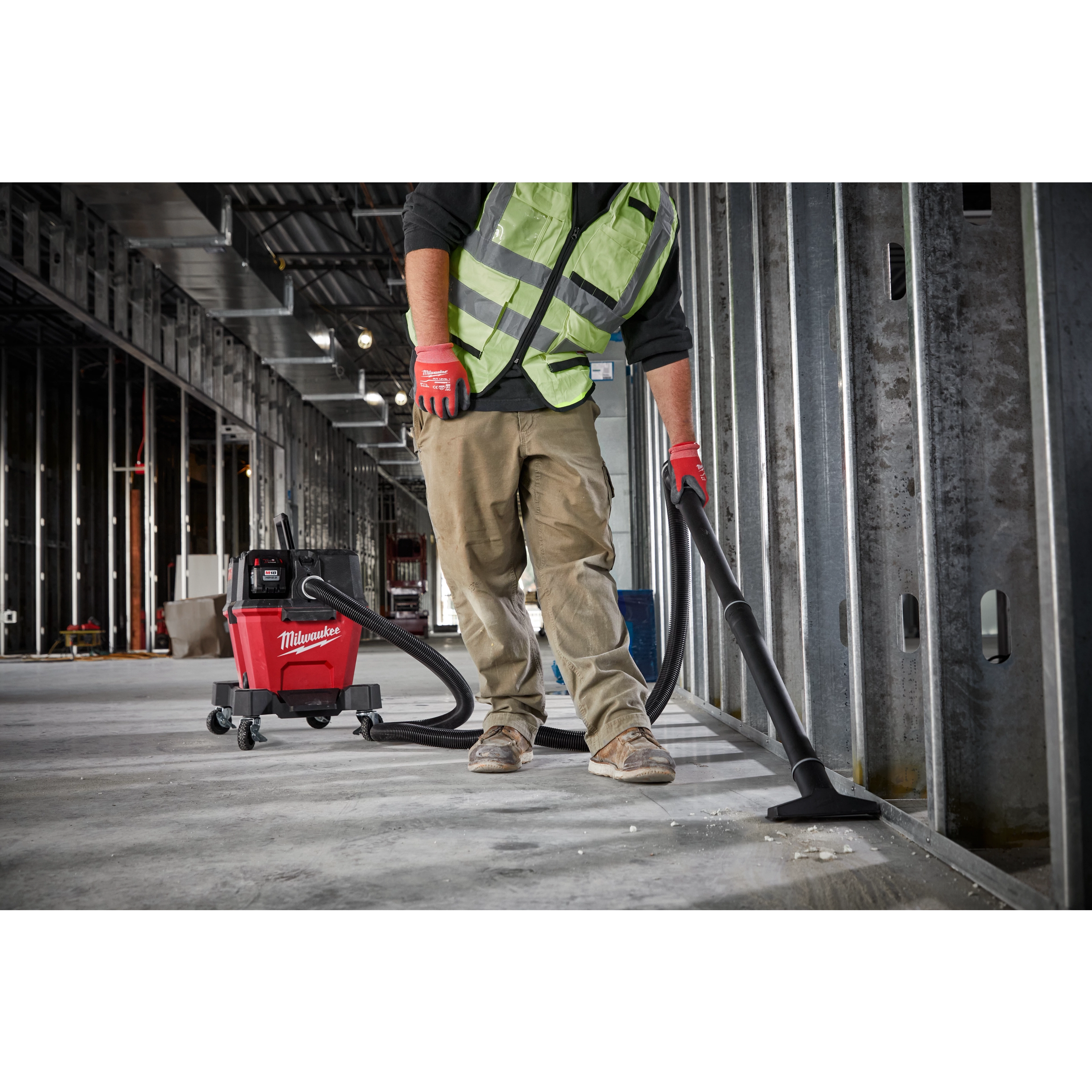 A worker in a green safety vest and red gloves uses a Milwaukee M18 FUEL 6 Gallon Wet/Dry Vacuum to clean a construction site.