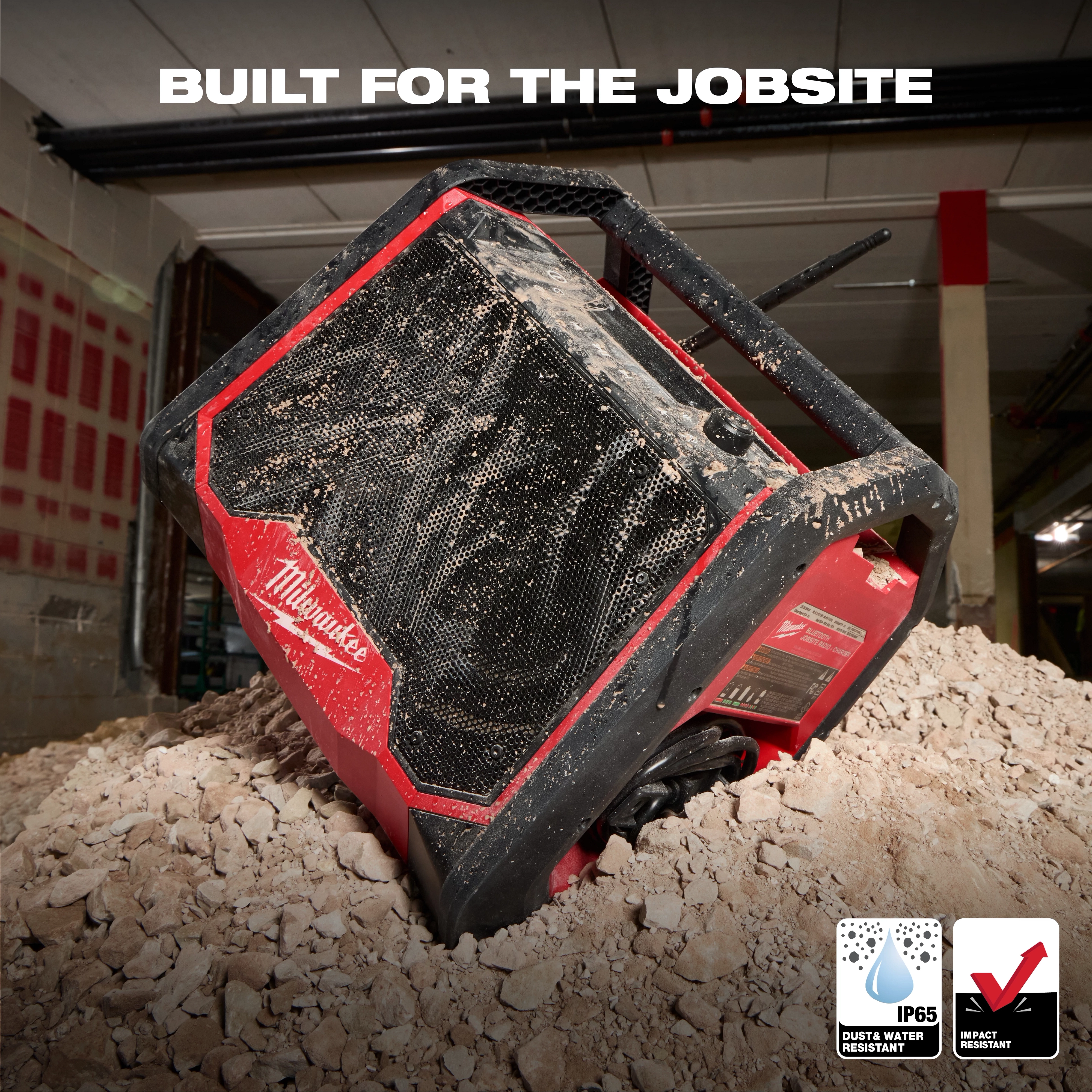 The M18 BLUETOOTH JOBSITE RADIO + CHARGER is shown on a dusty construction site. The rugged, red and black device features dust and water resistance (IP65) and impact resistance. The product is designed for durability and tough work environments, enhancing the job site audio experience while charging tools.