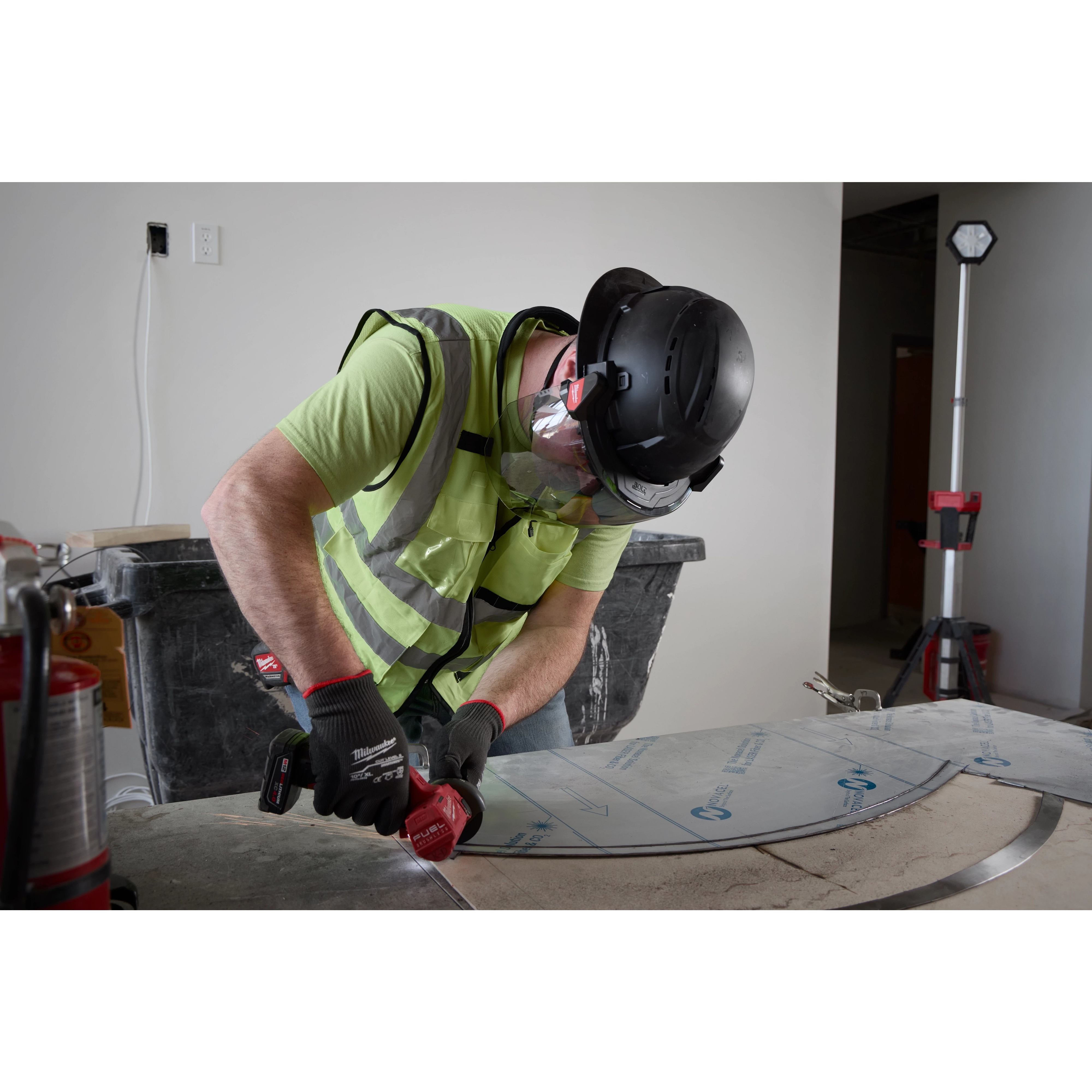 A worker wearing a BOLT™ Black Full Brim Vented Safety Helmet – Type 2, Class C, a high-visibility safety vest, safety gloves, and protective eyewear uses a power tool to work on a metal surface in a construction environment.
