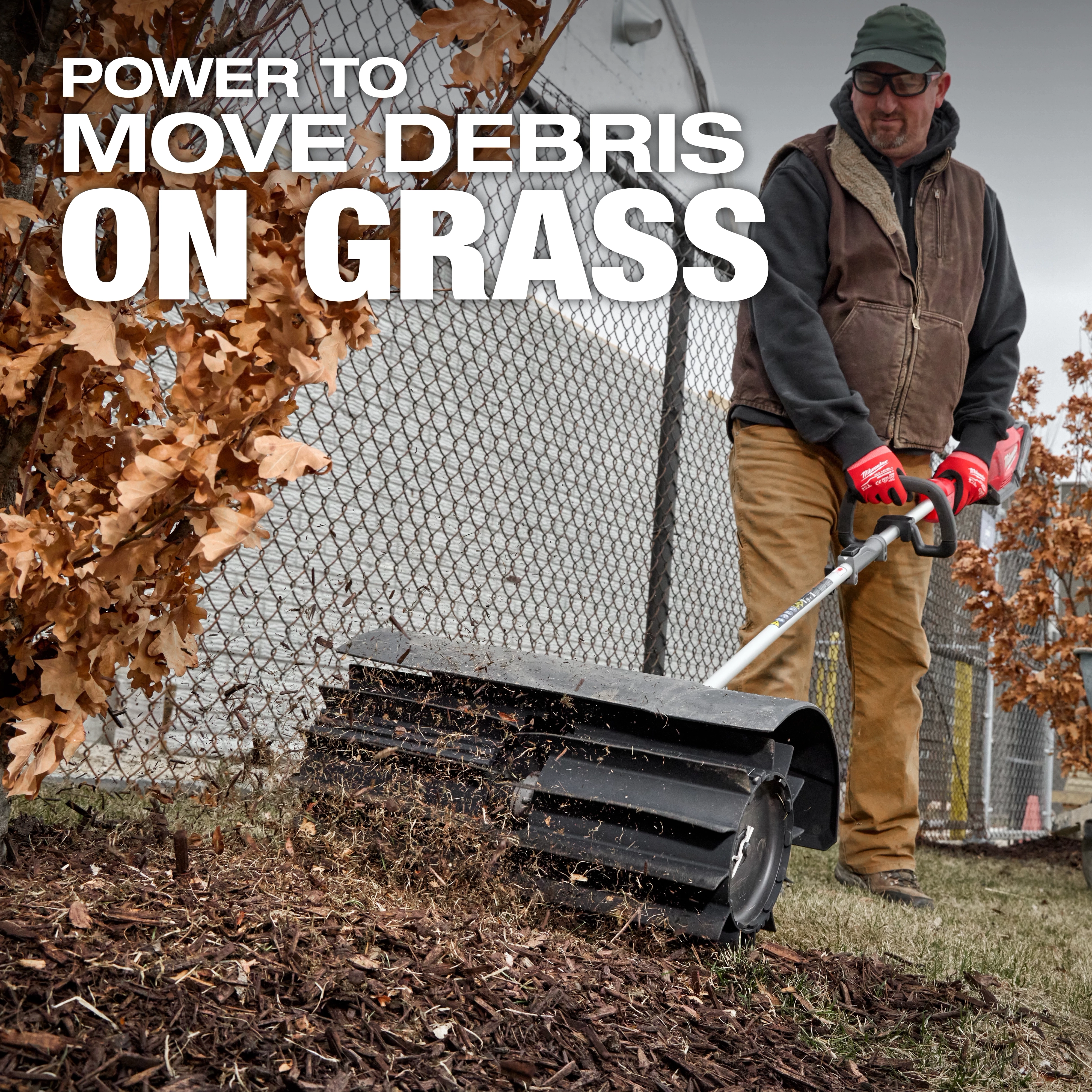A person using a power sweeper to clear debris from grass near a fence and trees. The text "Power to move debris on grass" is displayed at the top of the image. The person wears work gloves, a brown vest, and tan pants.