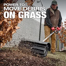 A person using a power sweeper to clear debris from grass near a fence and trees. The text "Power to move debris on grass" is displayed at the top of the image. The person wears work gloves, a brown vest, and tan pants.