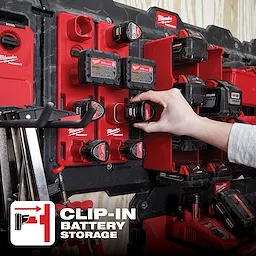 Hand places a battery in a PACKOUT M12 Battery Rack on a wall displaying multiple red battery packs and battery storage units.