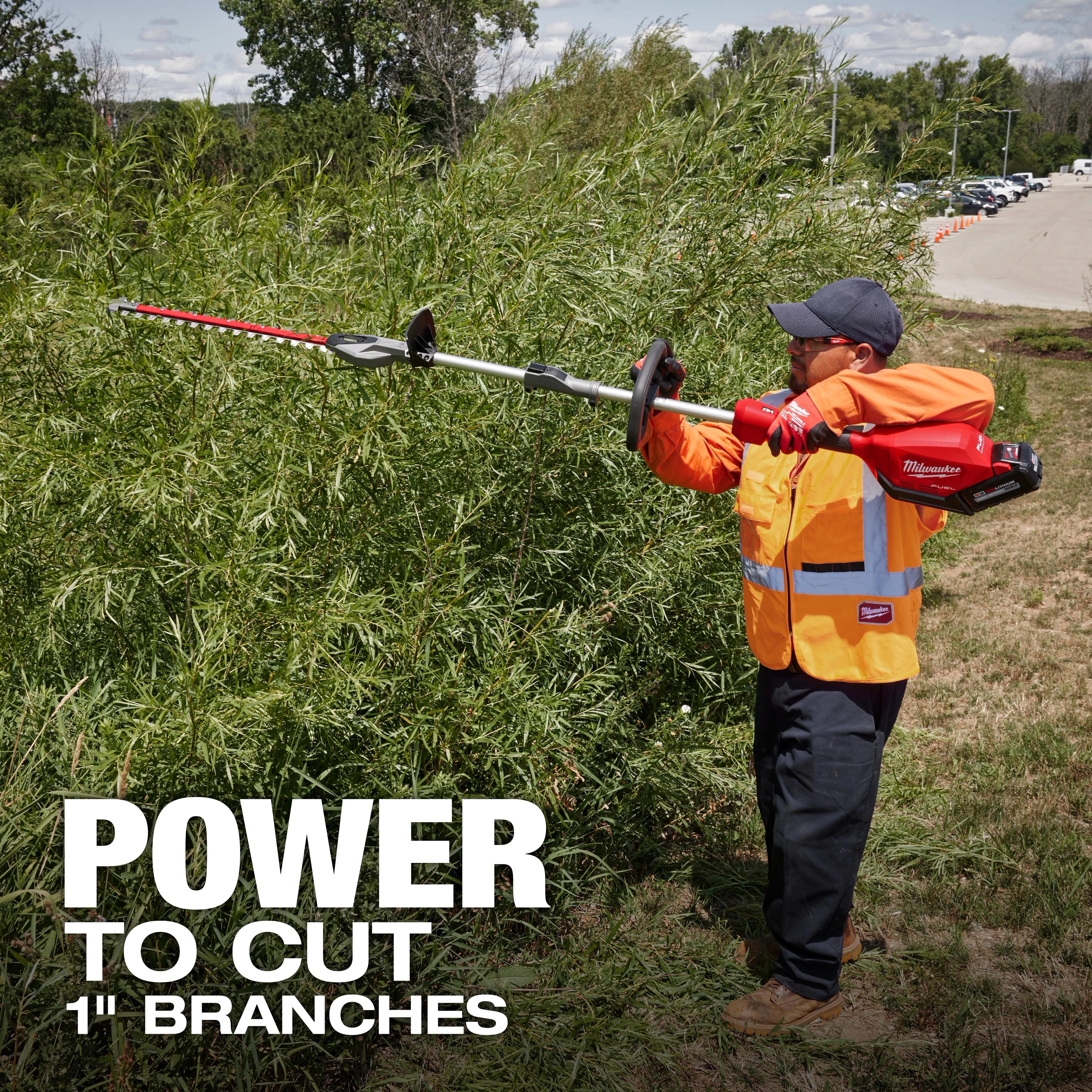 A person in an orange safety vest uses a Milwaukee power tool to trim large bushes. The text on the image reads "Power to cut 1" branches." Trees and a parking lot are in the background.