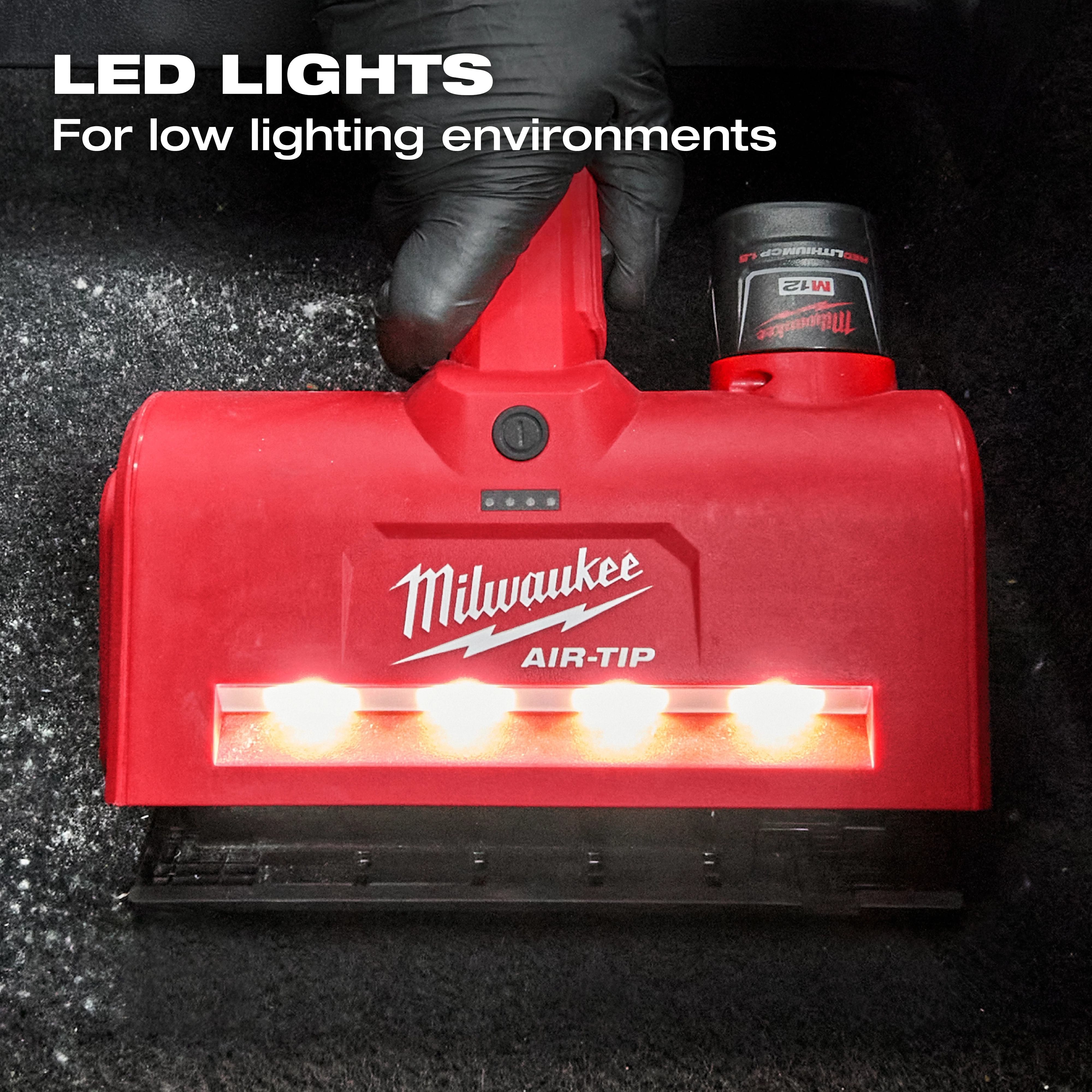 A red M12™ AIR-TIP™ Utility Nozzle with bright LED lights for low lighting environments. The nozzle has a Milwaukee logo and is being held by a gloved hand. The device is shown in use on a dark carpeted surface.