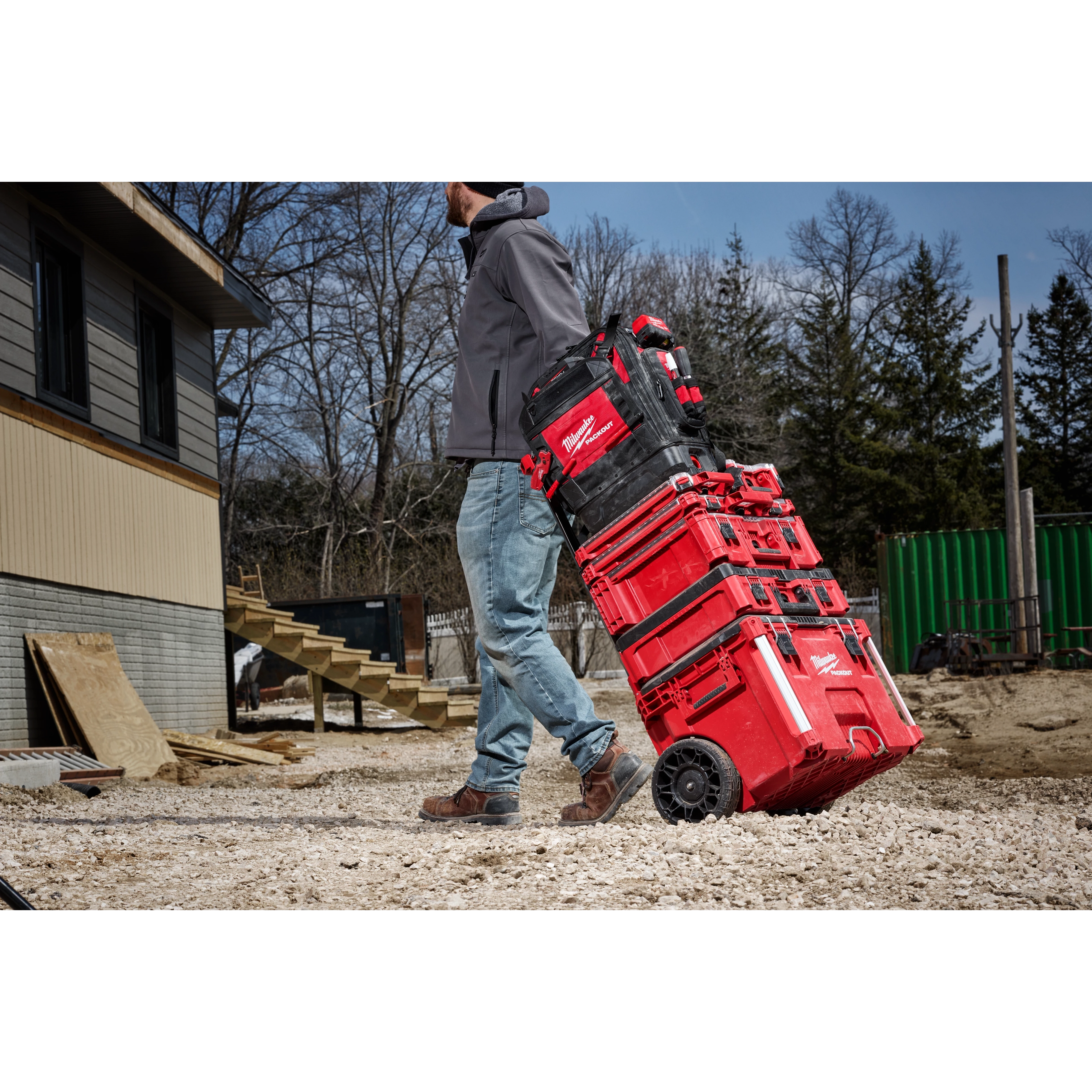 A person pulls a stack of red and black PACKOUT™ Structured Backpack and toolboxes on a wheeled cart at a construction site. The ground is covered in gravel, and there are trees and a building in the background.