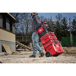 A person pulls a stack of red and black PACKOUT™ Structured Backpack and toolboxes on a wheeled cart at a construction site. The ground is covered in gravel, and there are trees and a building in the background.