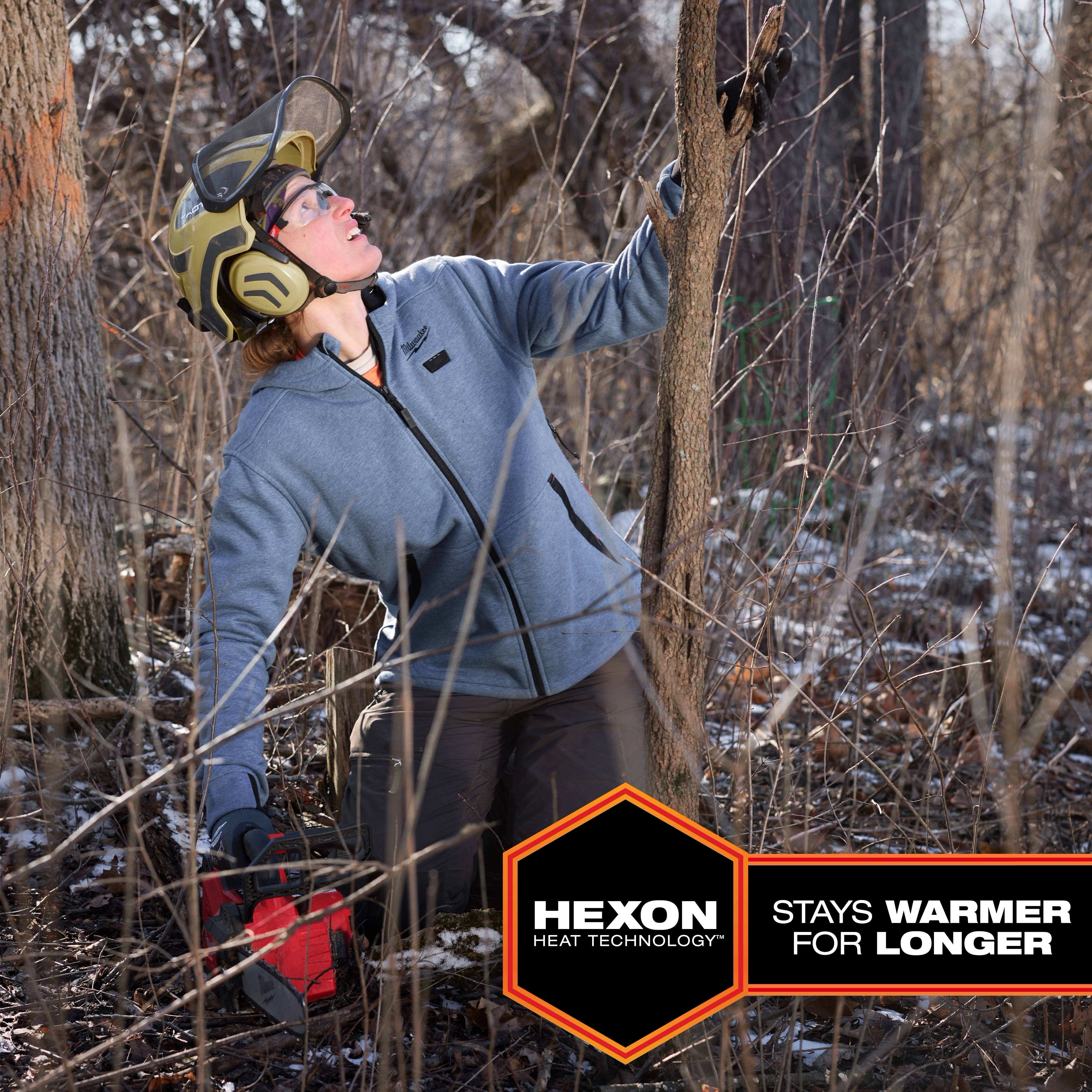 A person wearing protective gear works in a forest while wearing the M12™ Heated Hoodie. The hoodie is gray with black zippers and trim, featuring Hexon heat technology. A hexagonal badge on the image reads “Hexon Heat Technology™ stays warmer for longer.”