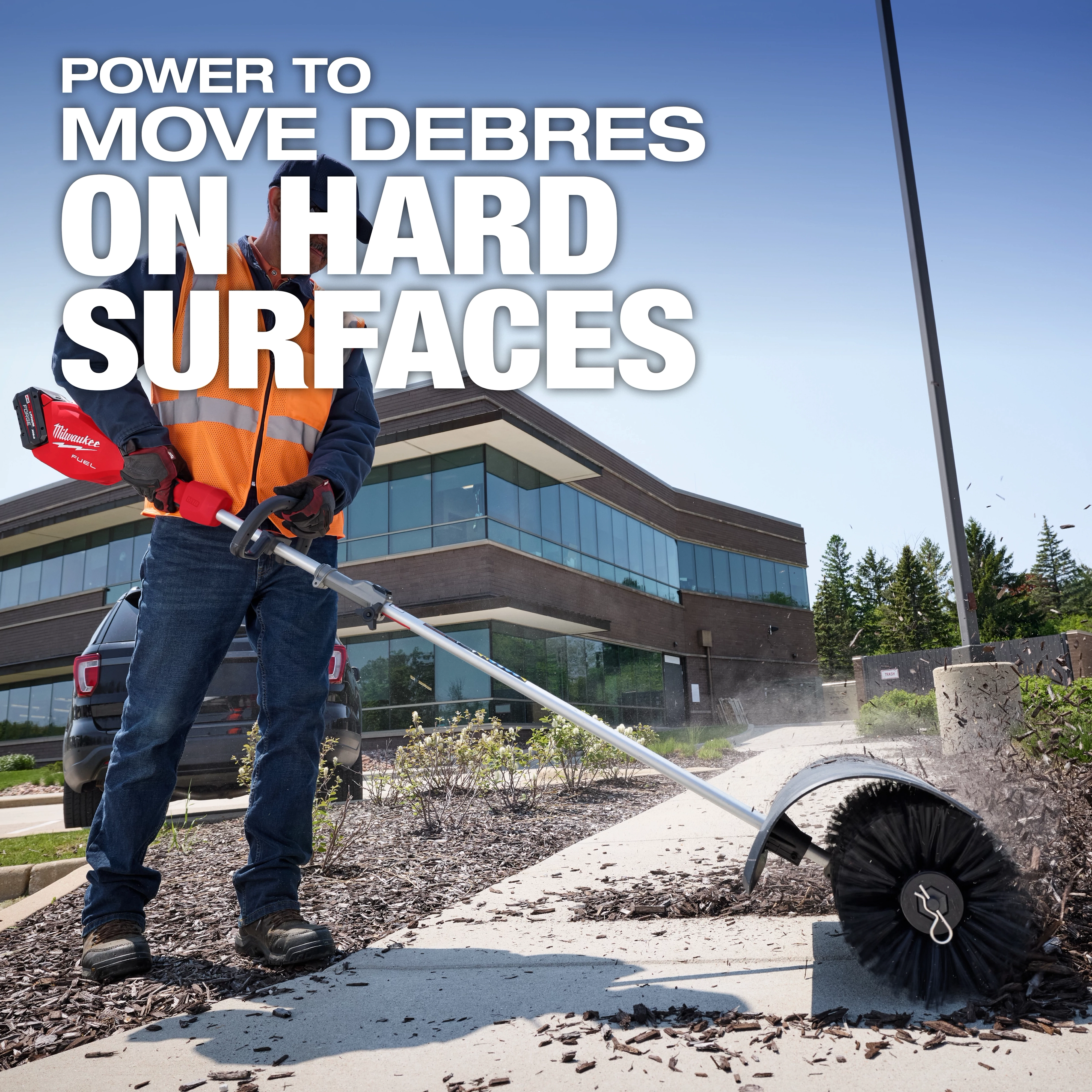 A worker wearing a safety vest uses a Milwaukee electric power tool with a rotating brush attachment to clear debris from a pathway outside a modern office building. Text on the image reads, "Power to move debris on hard surfaces."