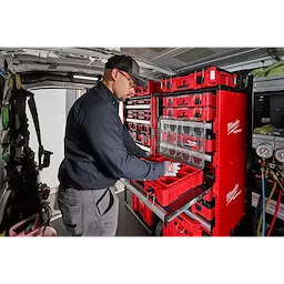 A worker organizes tools using the PACKOUT™ Rack Kit installed in a van. The red storage system includes multiple shelves and compartments, holding various Milwaukee toolboxes and organizers, ensuring efficient storage and easy access to tools.