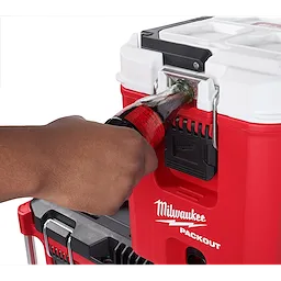 A person is using a built-in bottle opener on a red and white PACKOUT™ 16QT Compact Cooler. The cooler has the Milwaukee logo on the front and is stacked on another PACKOUT™ unit.