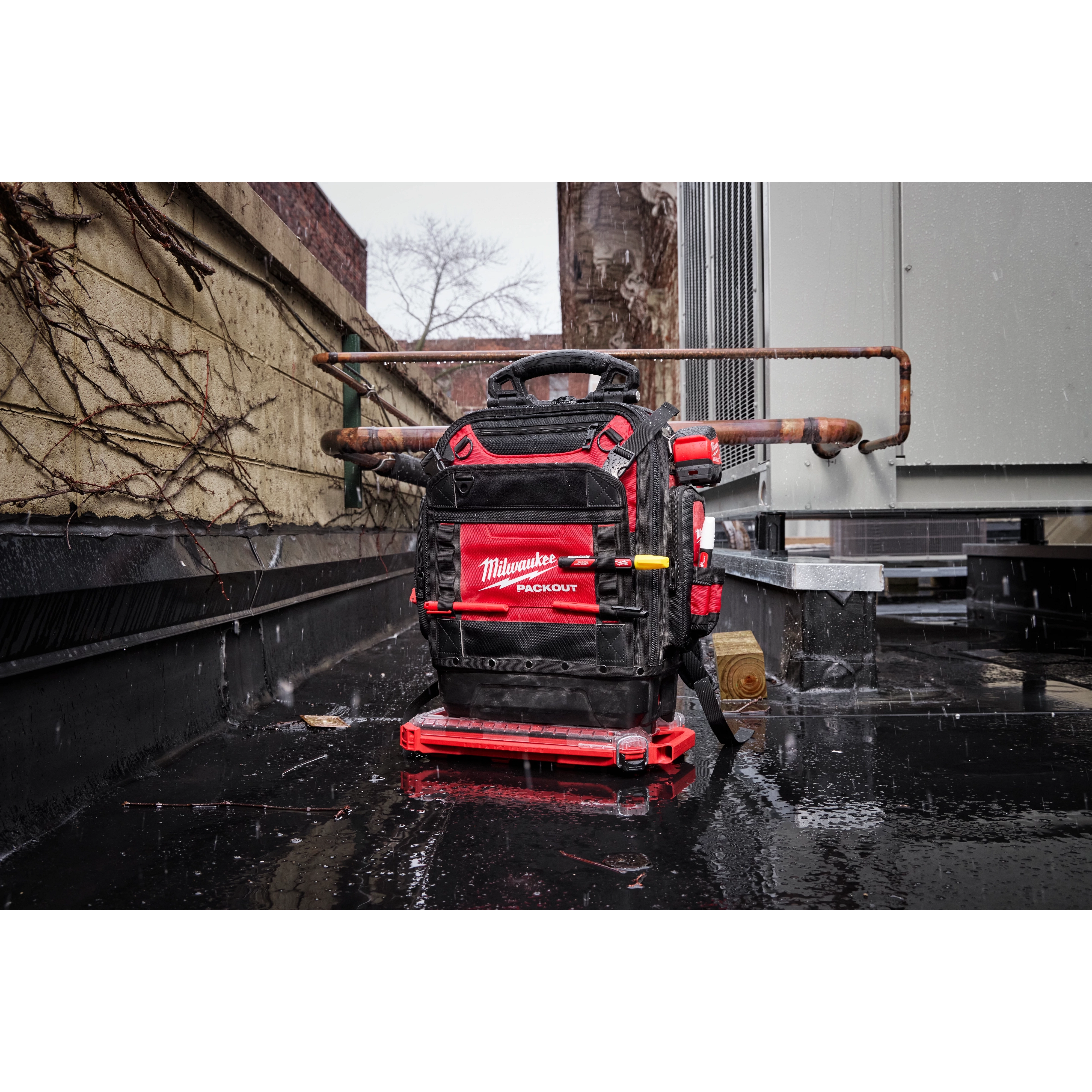 The PACKOUT™ Structured Backpack is shown in an industrial outdoor setting, resting on wet pavement. The black and red backpack features several compartments and the "Milwaukee" logo. It is designed for tools, with visible reinforcements and handles.
