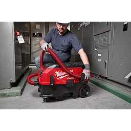 A person is using a Milwaukee vacuum cleaner connected to a red 10%27 Accessory Hose in an industrial setting. The vacuum cleaner is red and black with wheels for mobility. The background displays industrial equipment and control panels.